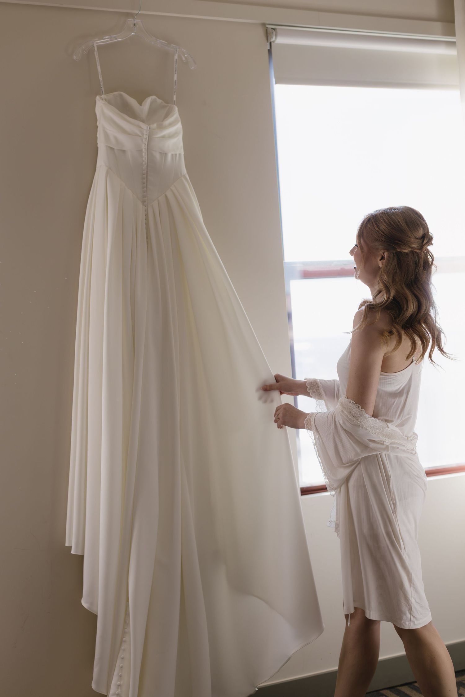 Series of shots showing woman in white robe examining wedding dress on hangers in sunlit room.