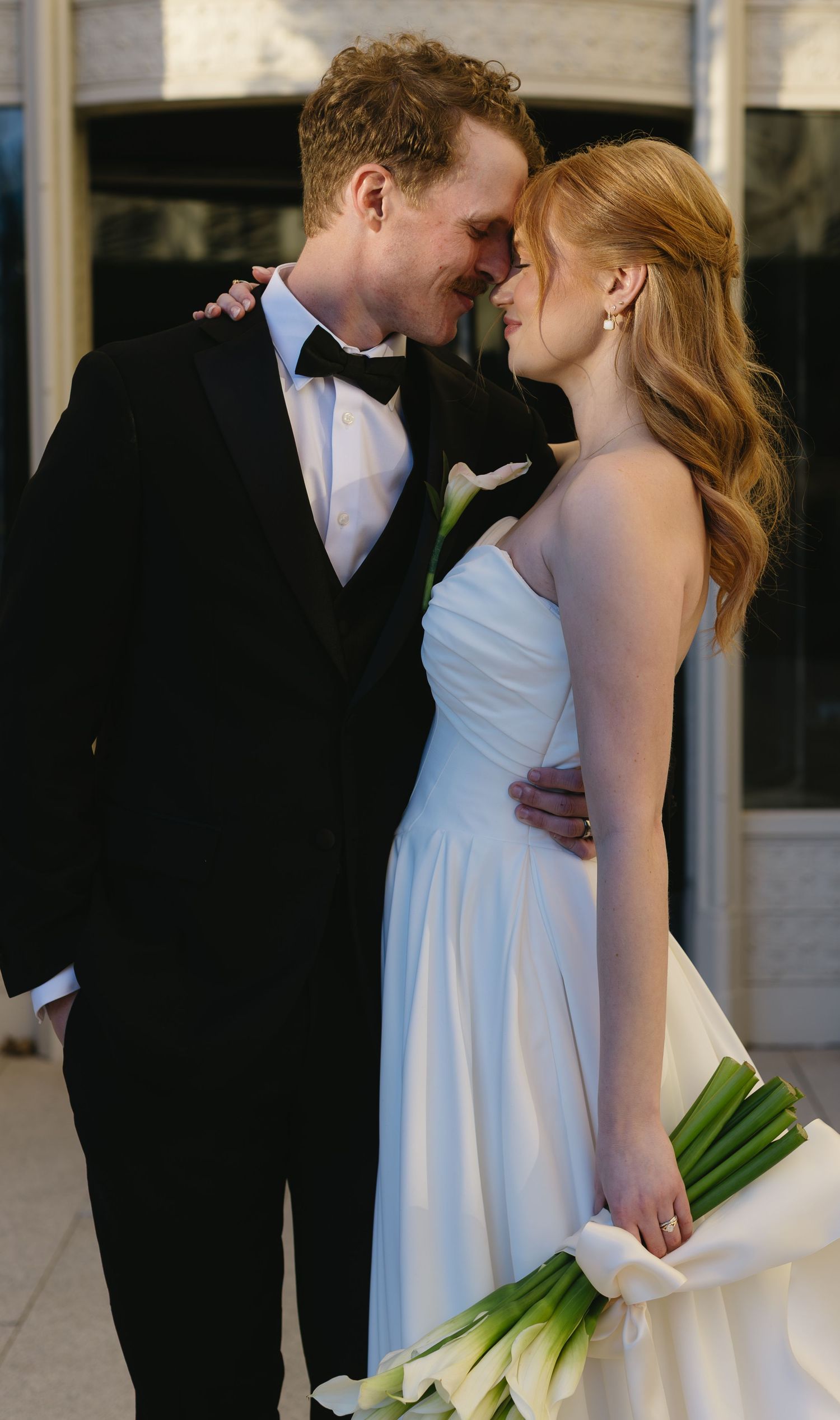 Intimate moment between newlyweds with white calla lily bouquet against stone wall.
