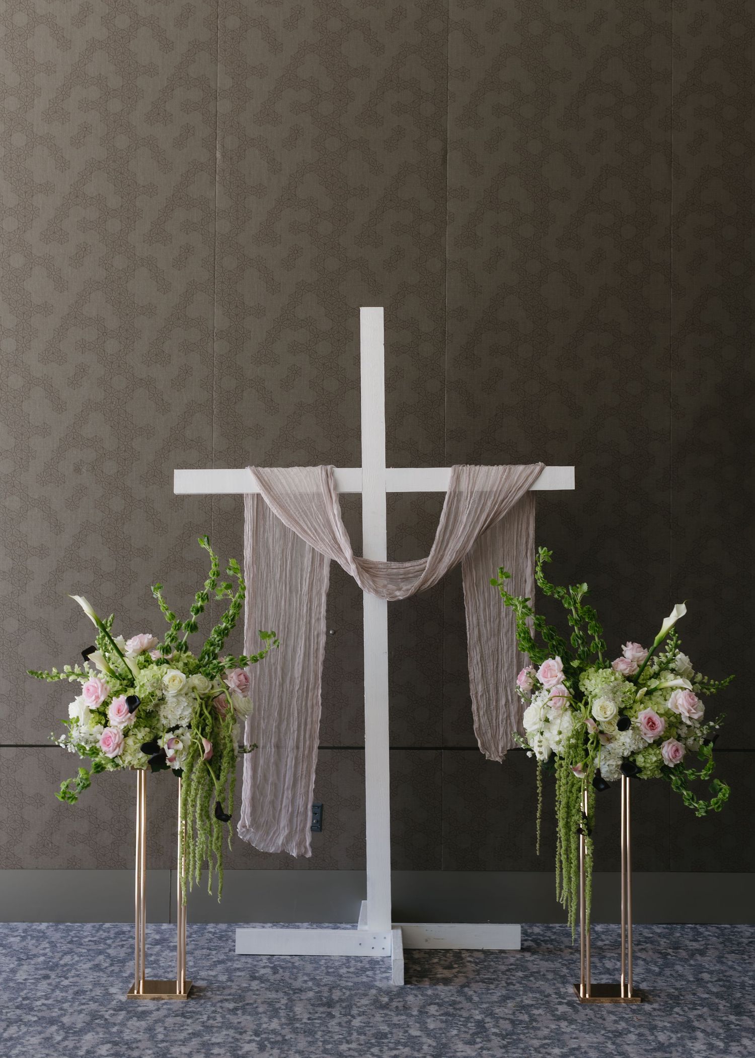 A white cross draped with fabric flanked by two tall floral arrangements in gold stands.