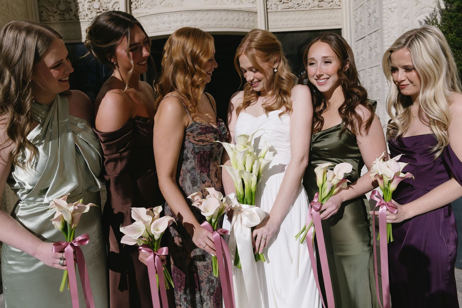 A bridal party in mixed jewel-toned dresses poses together holding white lily bouquets.