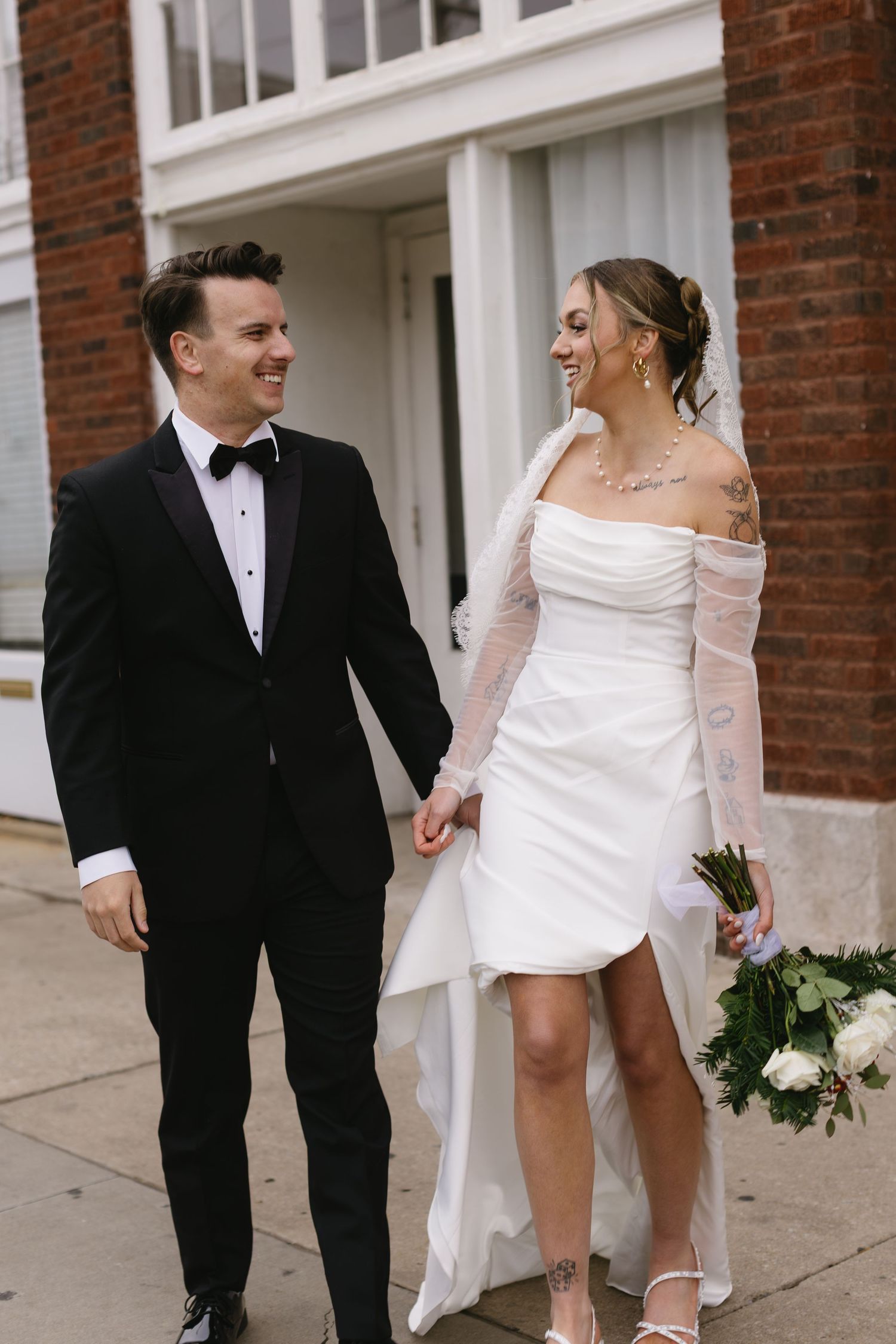 A newlywed couple shares a candid moment on a sidewalk with the bride holding a white bouquet.
