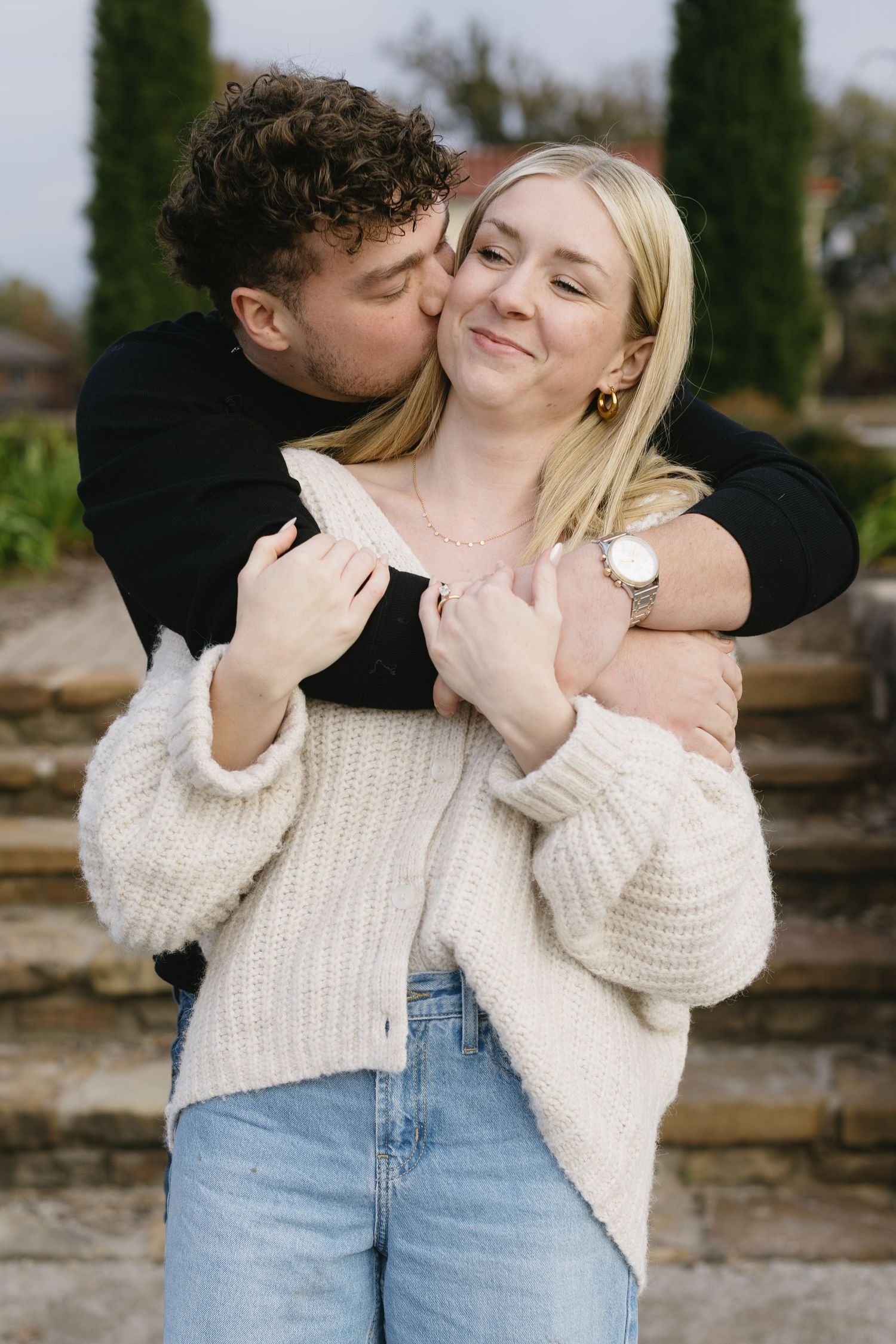 A romantic couple embraces outdoors wearing casual clothing of jeans and sweaters against a rustic wooden fence background.