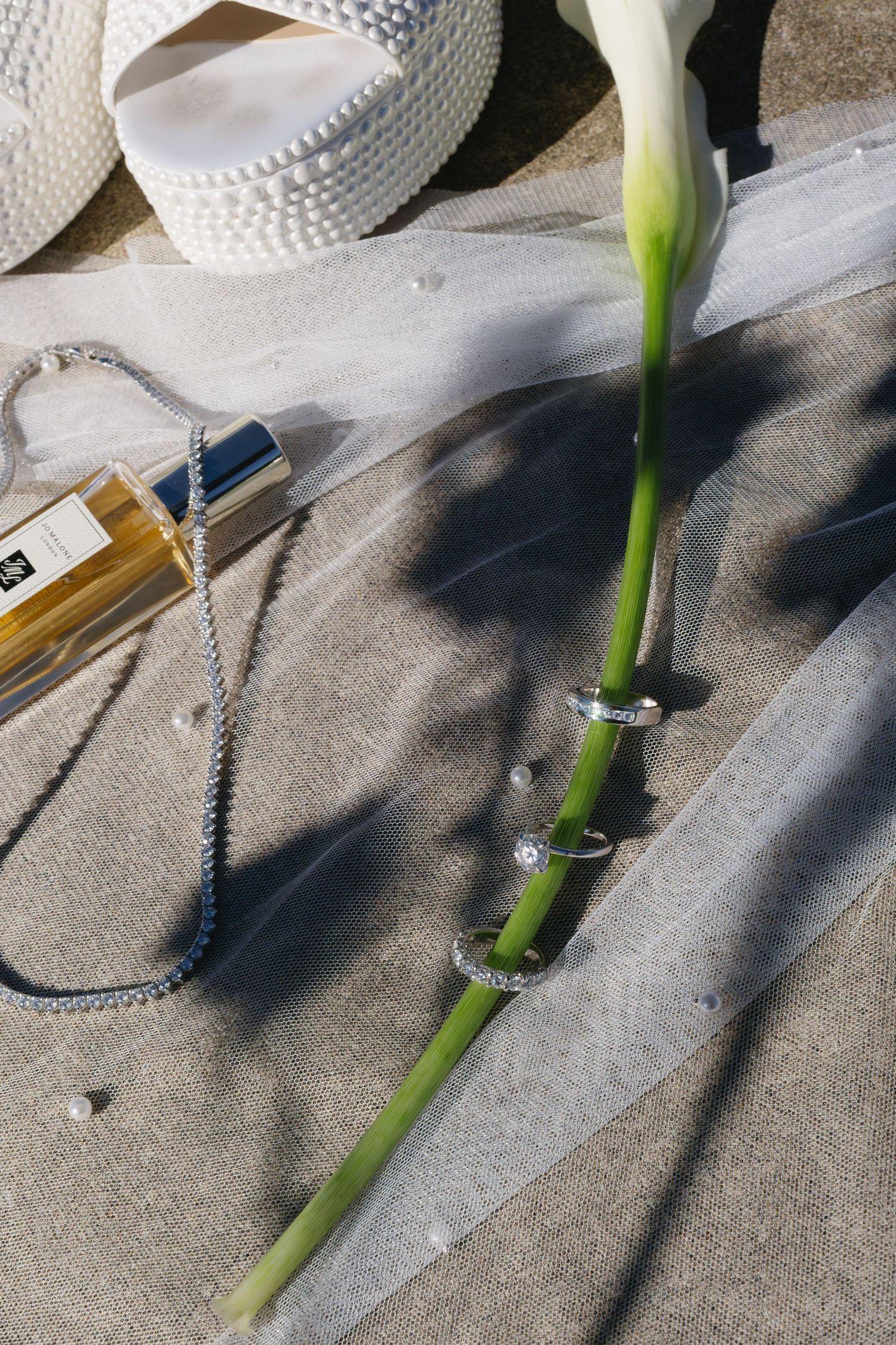 A white calla lily stem with jewelry and perfume bottle arranged on a textured surface.