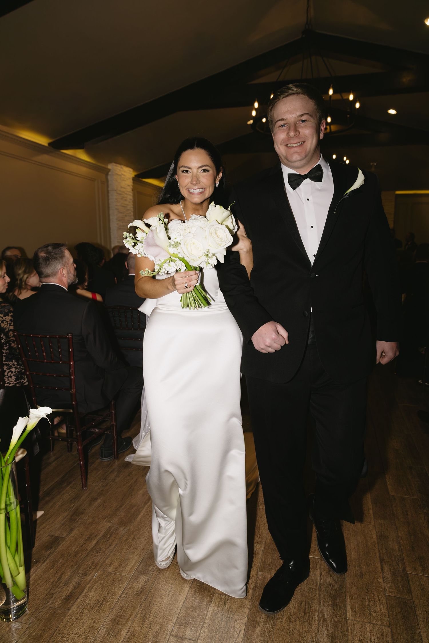 A newlywed couple walks arm in arm down a wooden floor with the bride carrying a white floral bouquet.