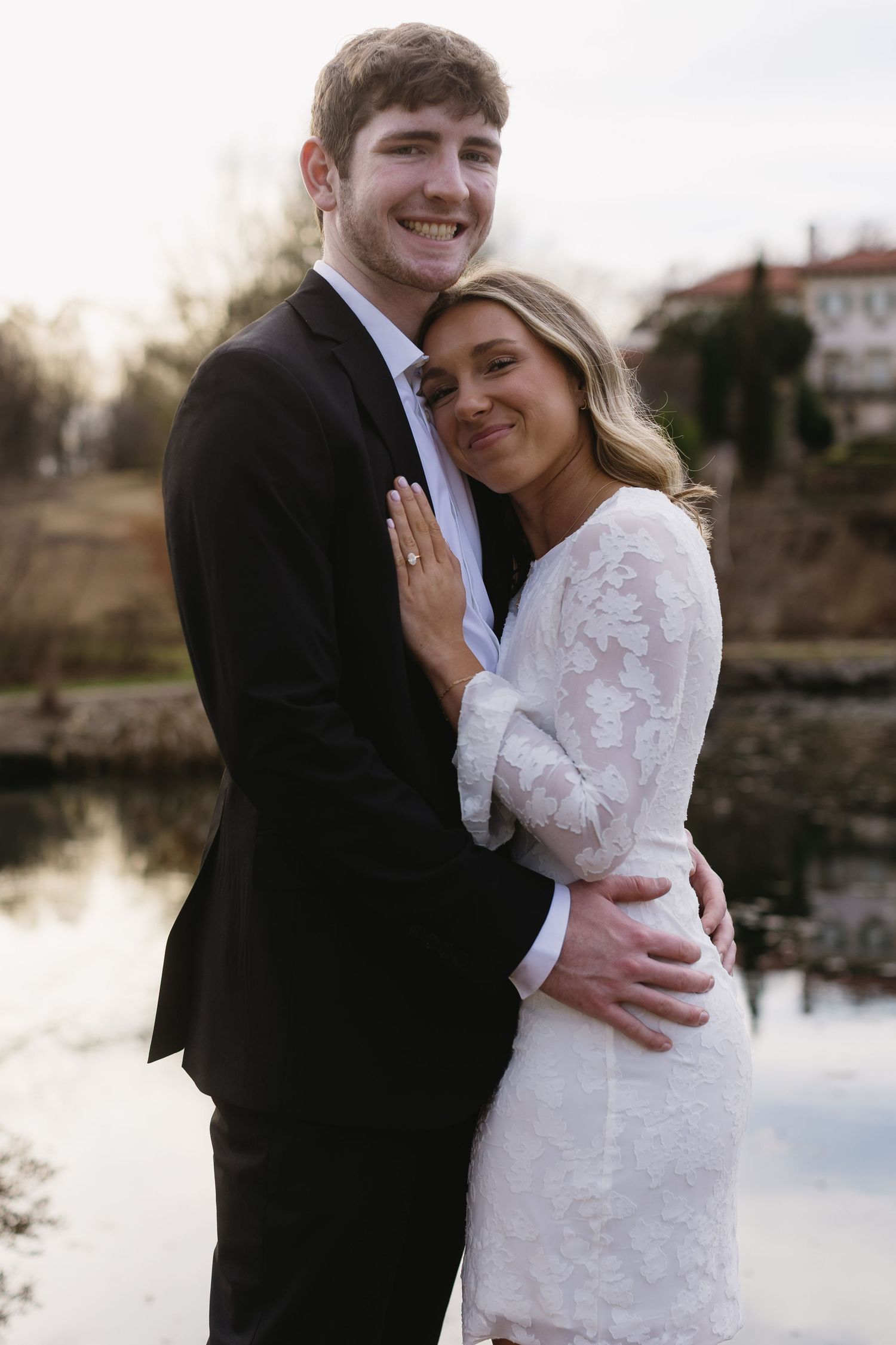 A couple in wedding attire embraces outdoors with water and buildings in the background.