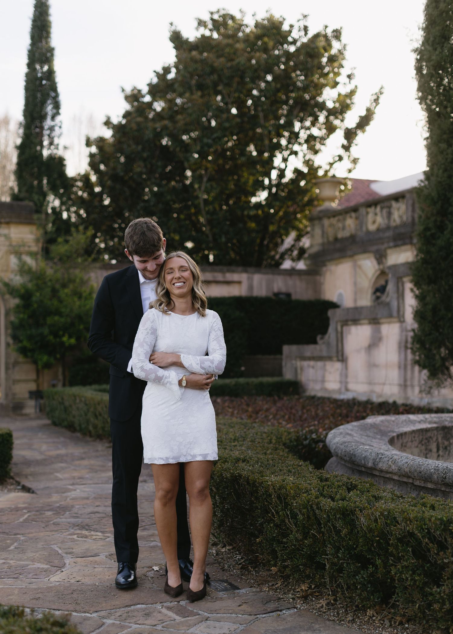 A couple shares a romantic moment among cypress trees and stone pathways.