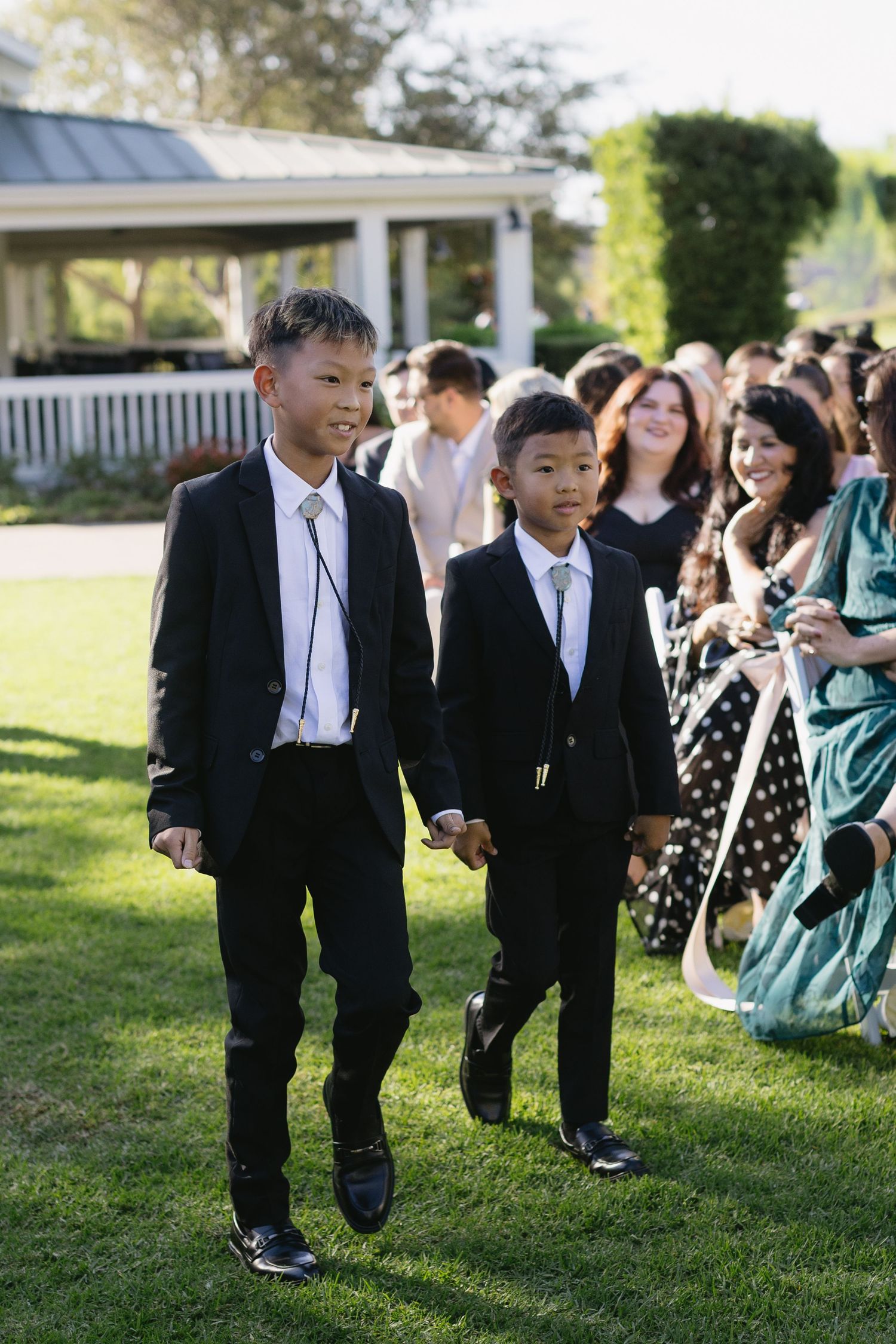 Young ring bearers in suits walking across sunny lawn during wedding ceremony.