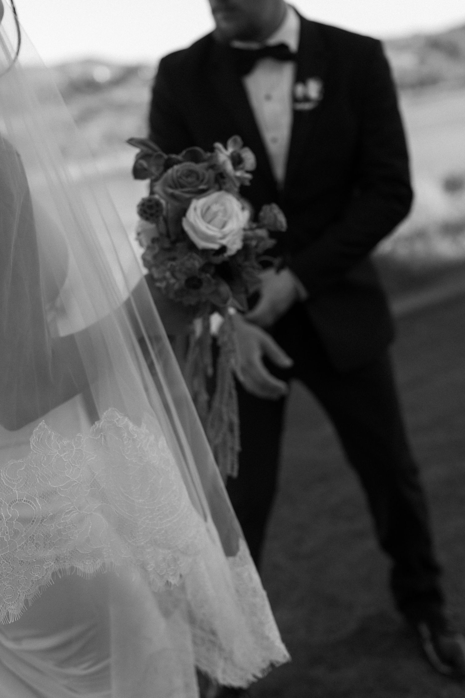 A black and white detail shot of a wedding bouquet with roses and a tuxedo in the background.