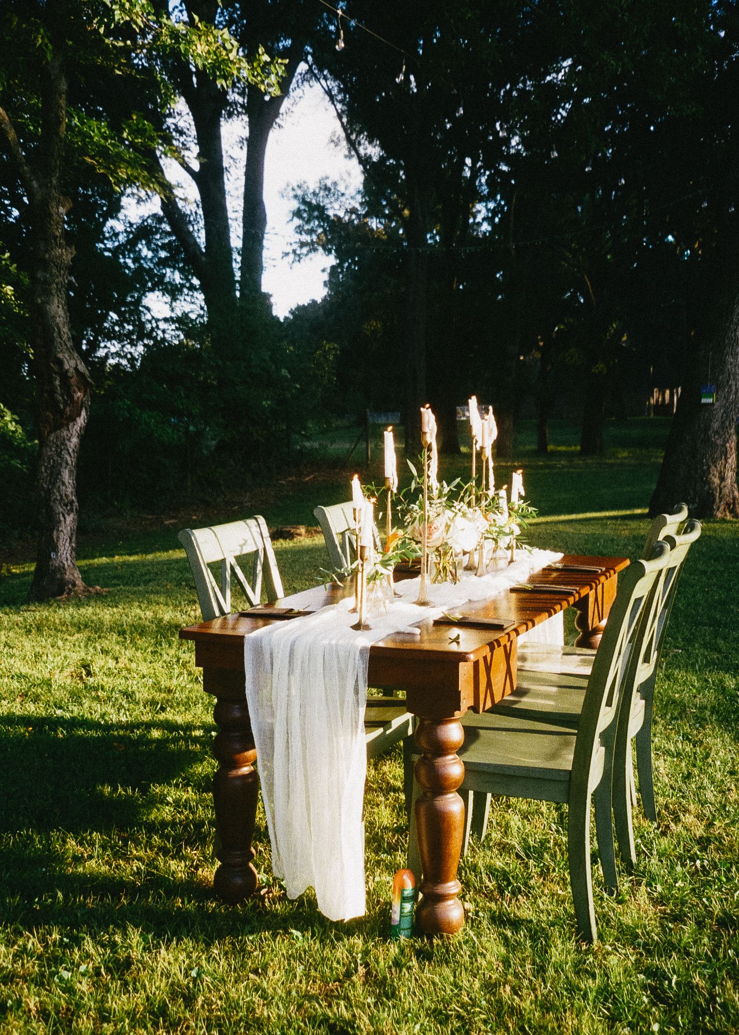 Long rustic wooden dining table with white linens and cross-back chairs set up outdoors for an elegant garden event.