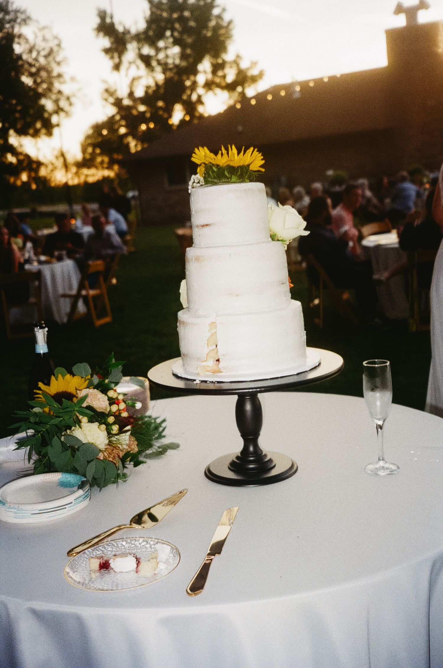 Three elegant white wedding cakes decorated with sunflowers and greenery sit on tables at an outdoor sunset reception.