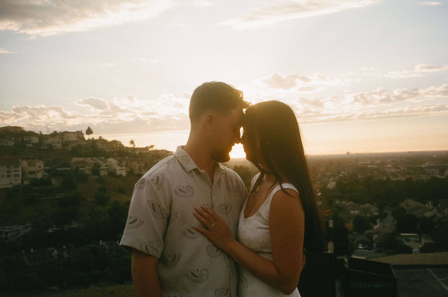 A couple shares a romantic moment at sunset against a coastal landscape backdrop.