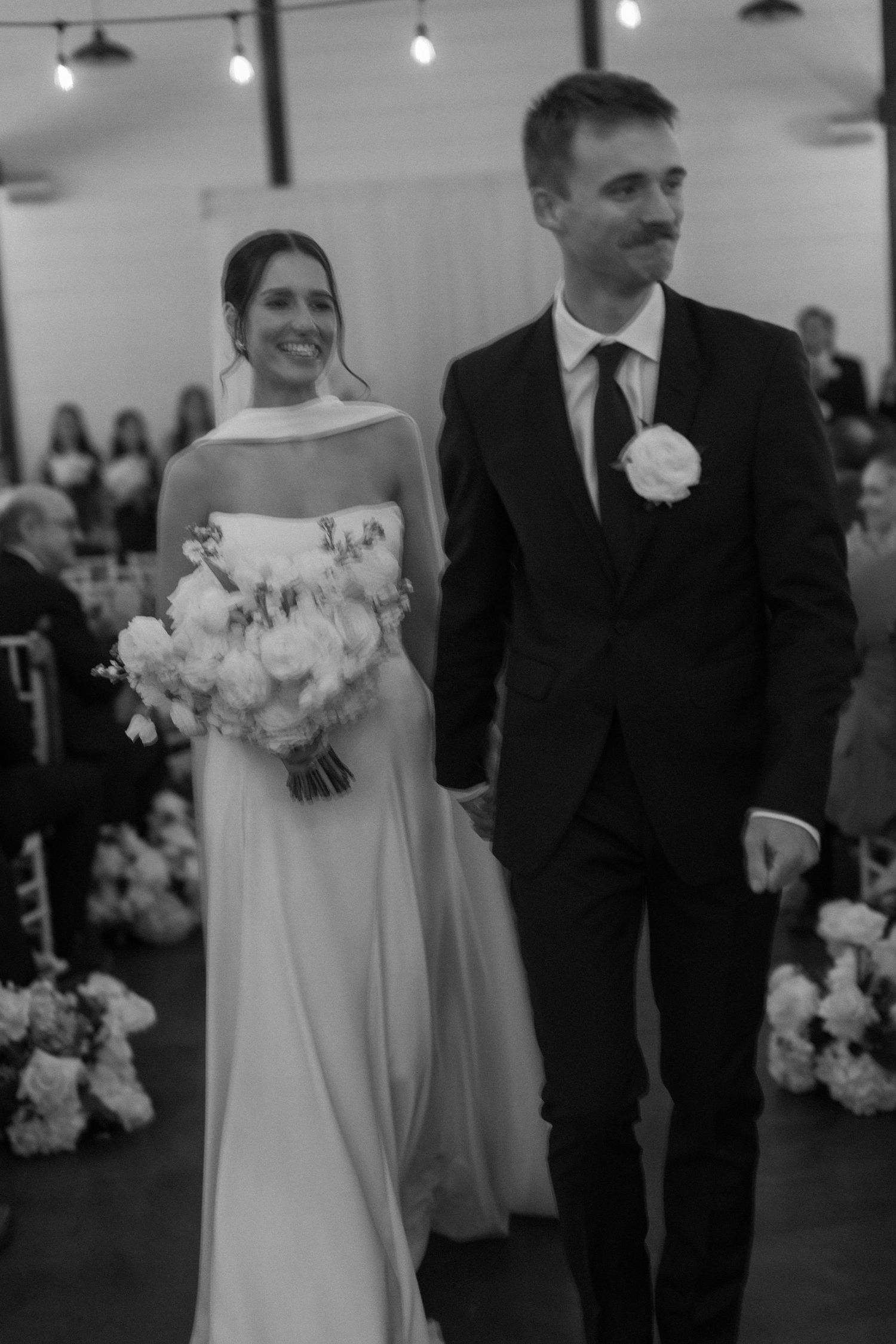 Indoor wedding ceremony with string lights overhead and bride in white gown holding floral bouquet.