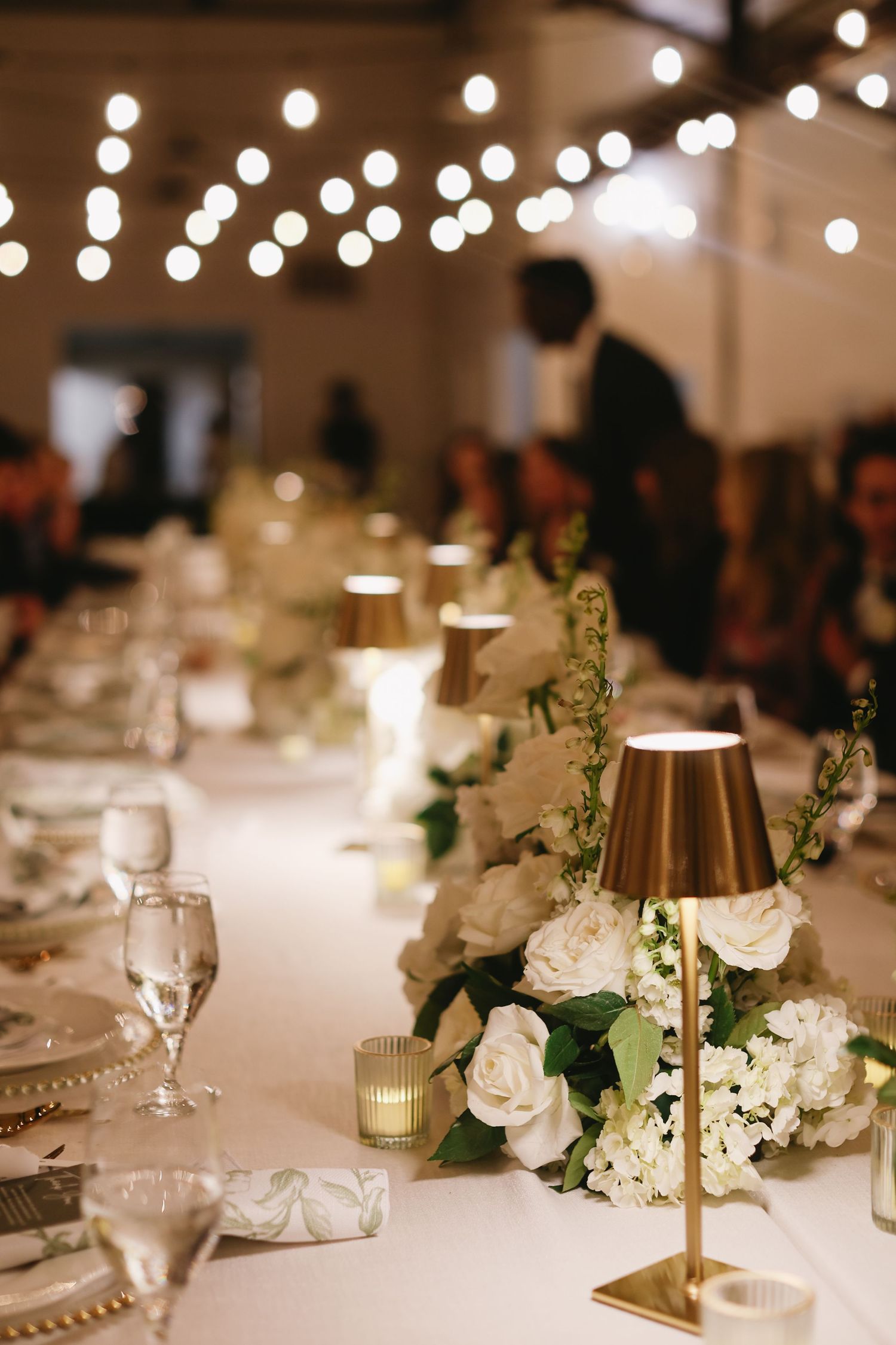White floral centerpieces and gold candle holders decorate wedding reception tables under ambient lighting.