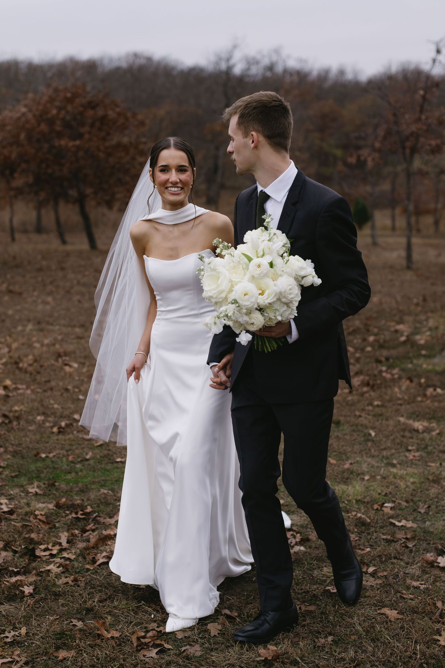 Elegant wedding couple walks together holding white bouquet in autumn orchard setting.