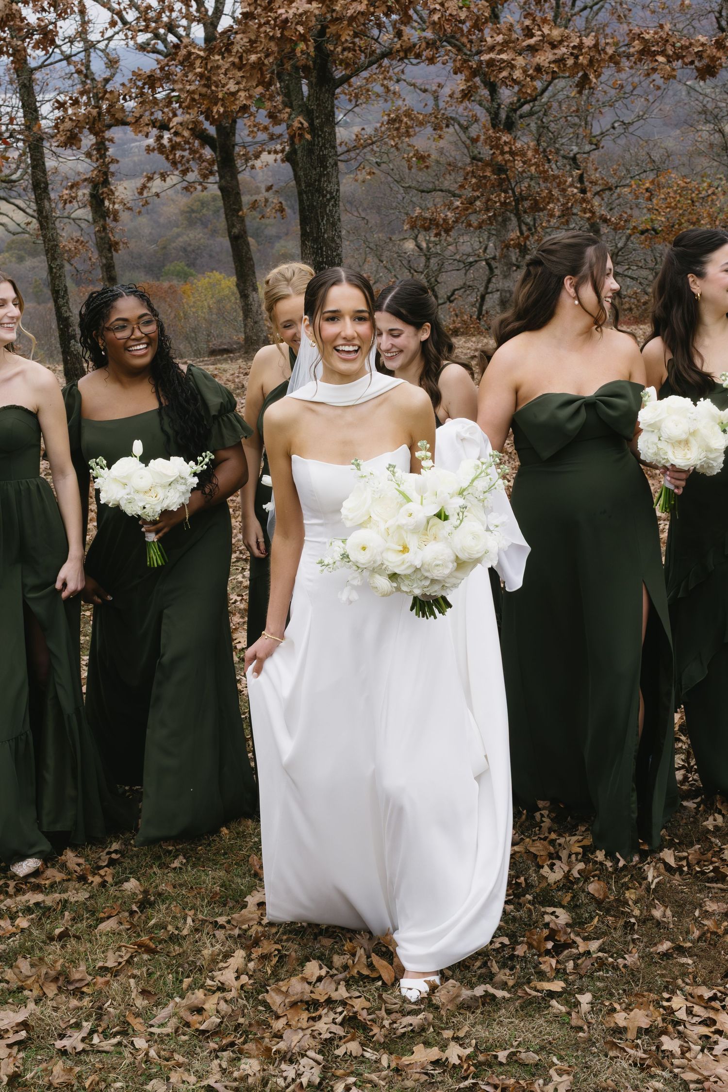Wedding party stands among fall foliage with bridesmaids in black dresses holding white bouquets.