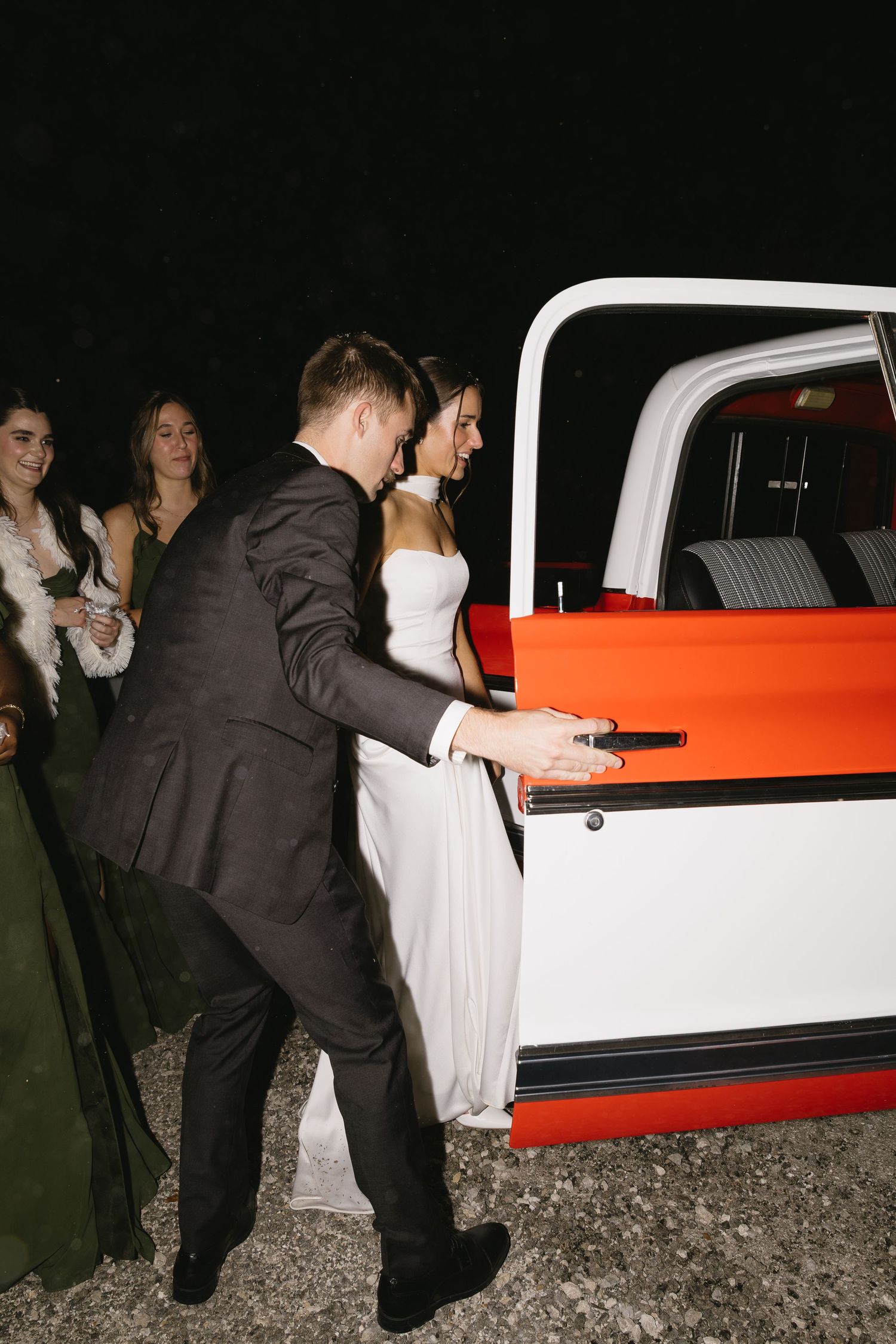 A couple in wedding attire approaches a white and orange vintage truck after their celebration.