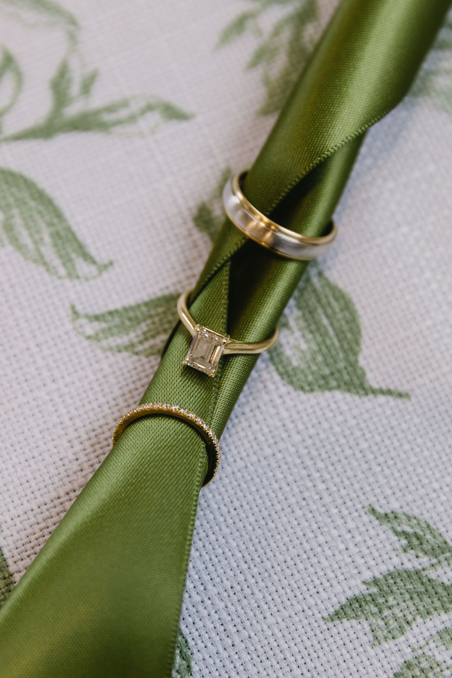 Wedding rings displayed on an olive green ribbon against a white and green floral patterned background.