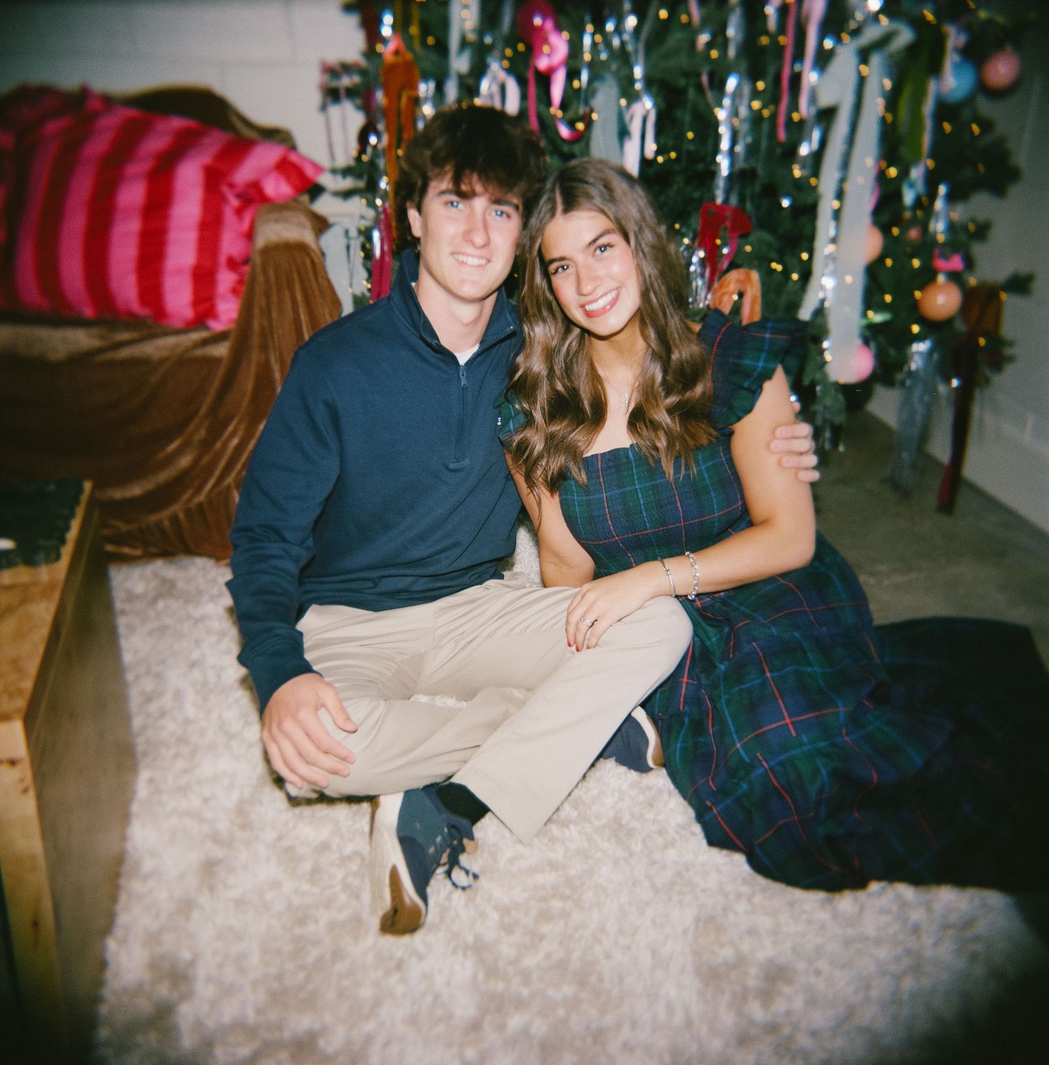 A couple sits together on a carpet in front of a decorated Christmas tree with festive holiday decor visible.