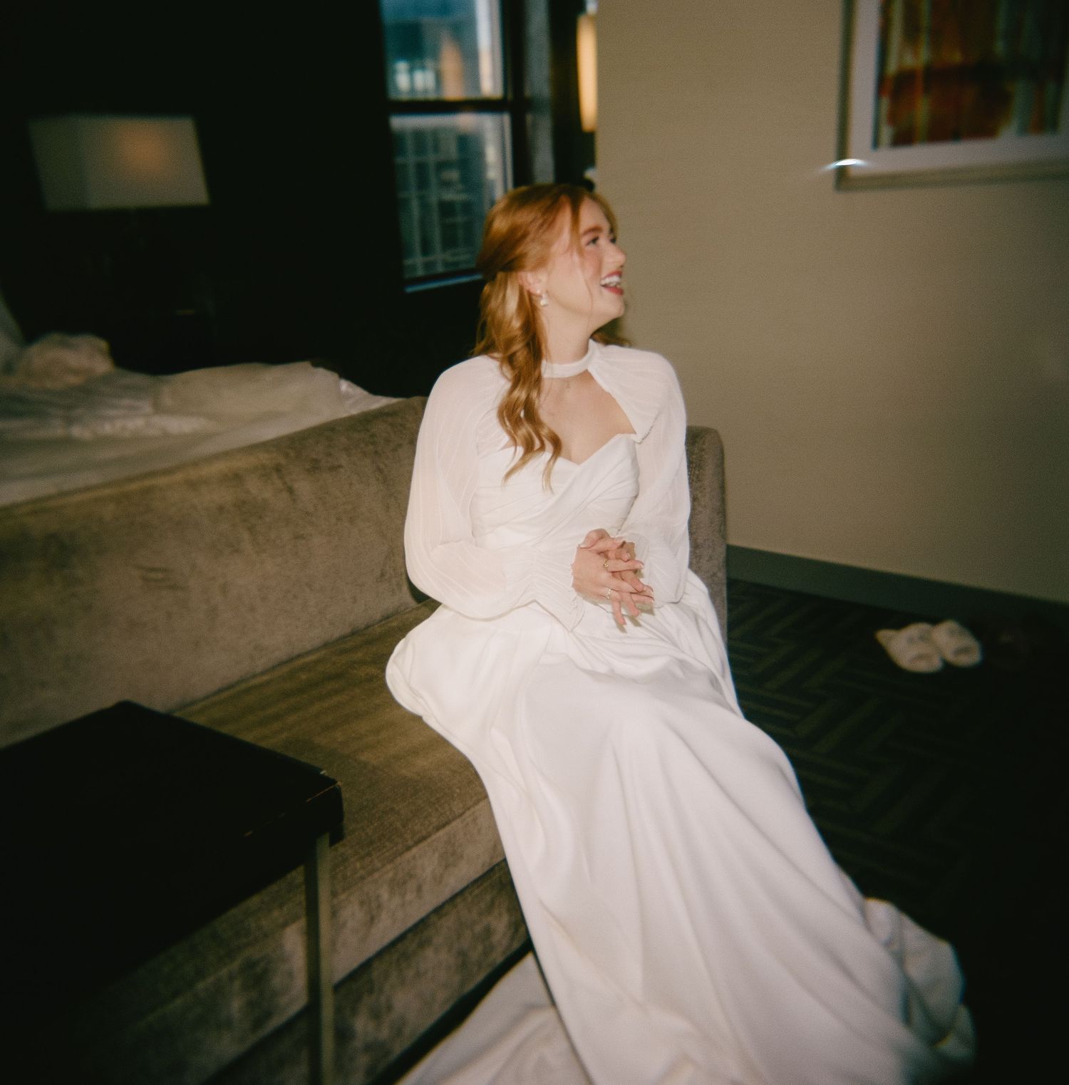Bride in flowing white dress reclining on dark furniture in intimate setting.