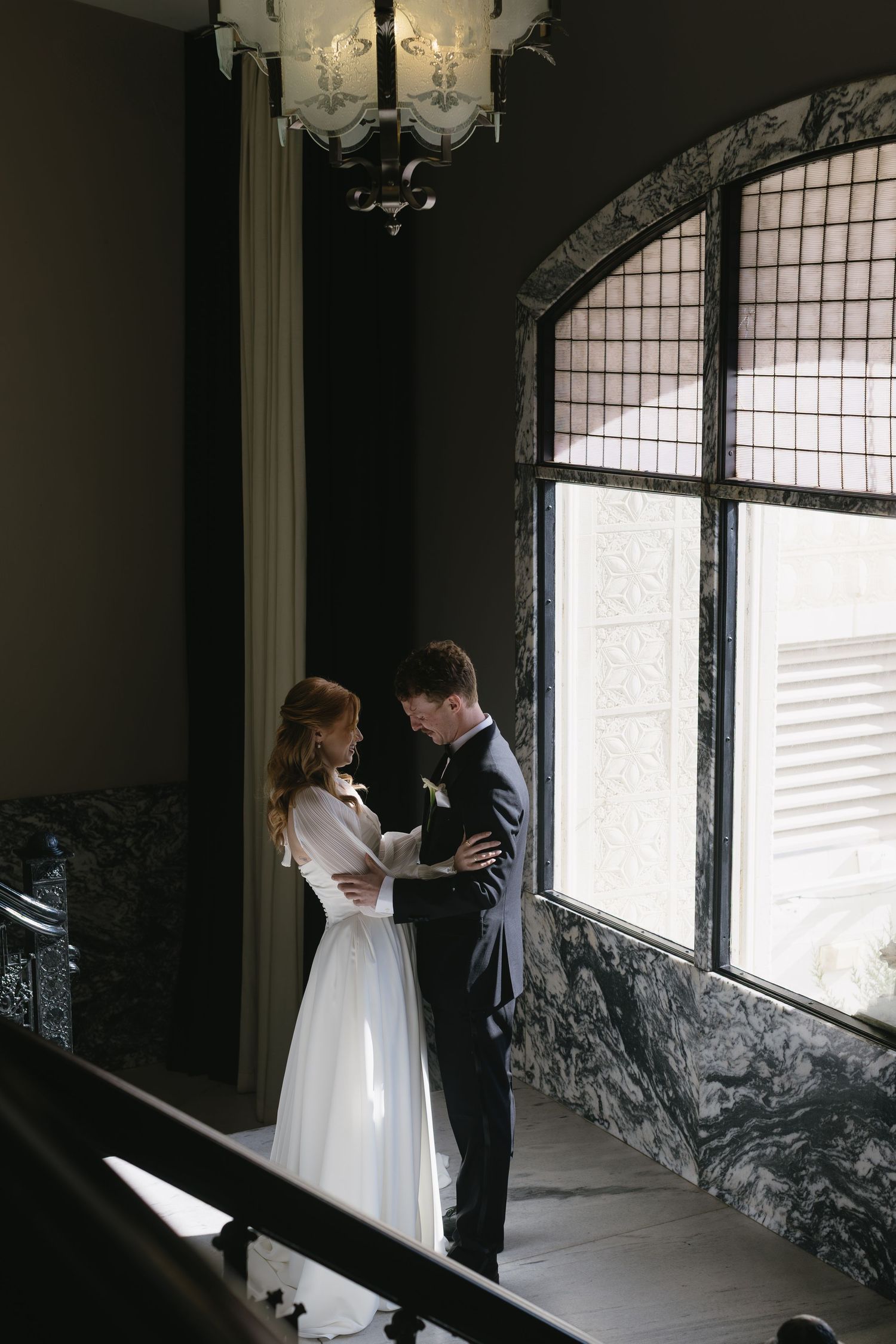 Wedding couple shares intimate moment by large arched windows with marble details.