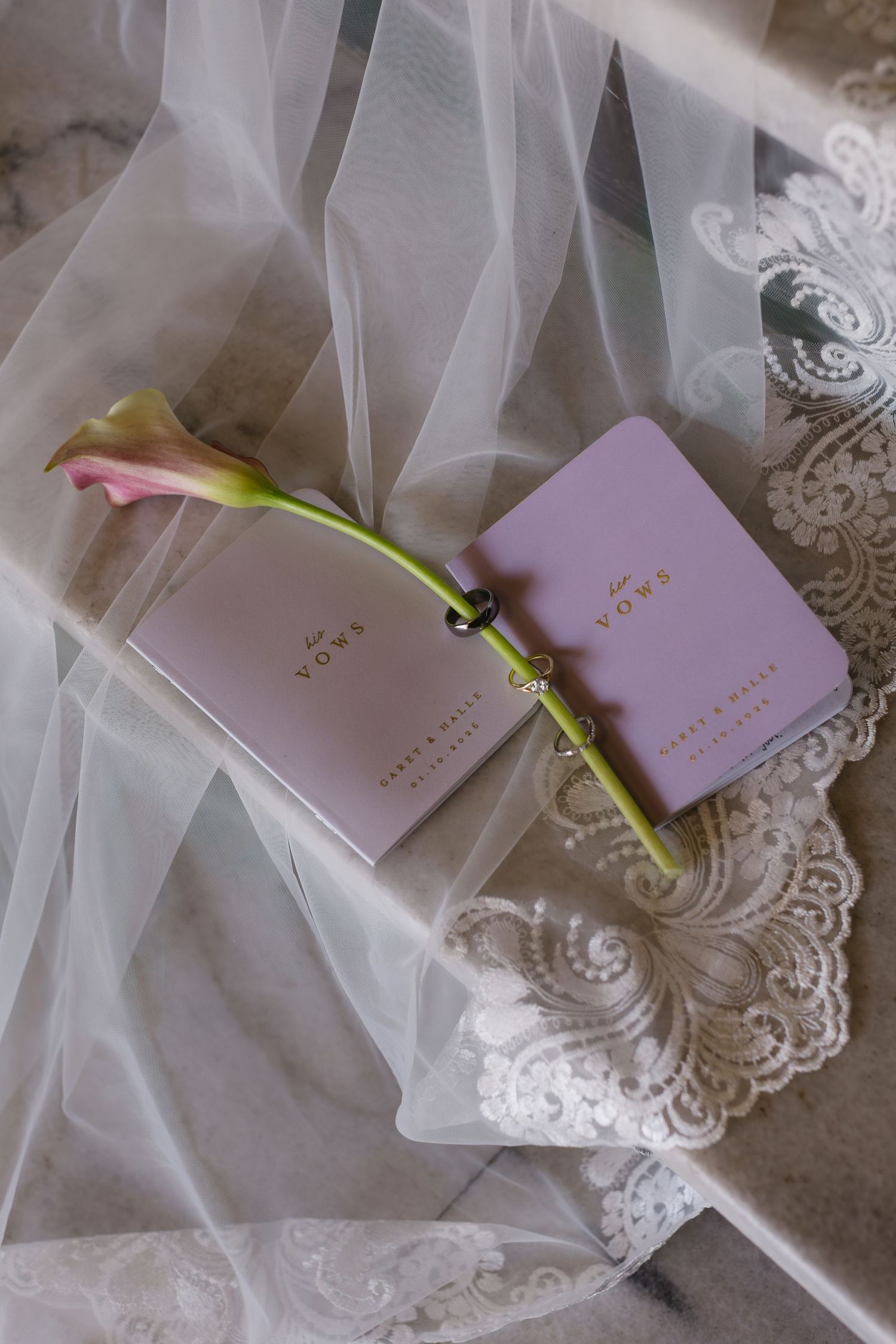 Pink wedding vow books rest on delicate white lace and tulle fabric with a single green stem.