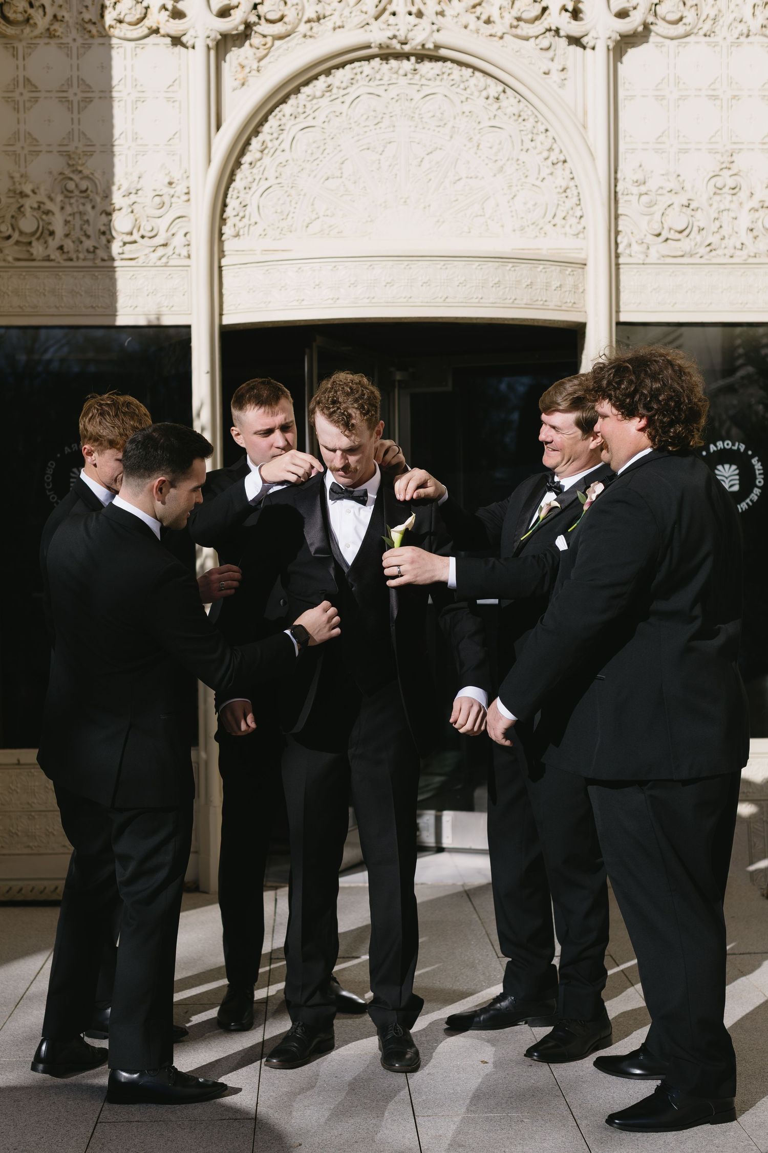 Groomsmen in black tuxedos line up against an ornate stone wall for a fun photo sequence.