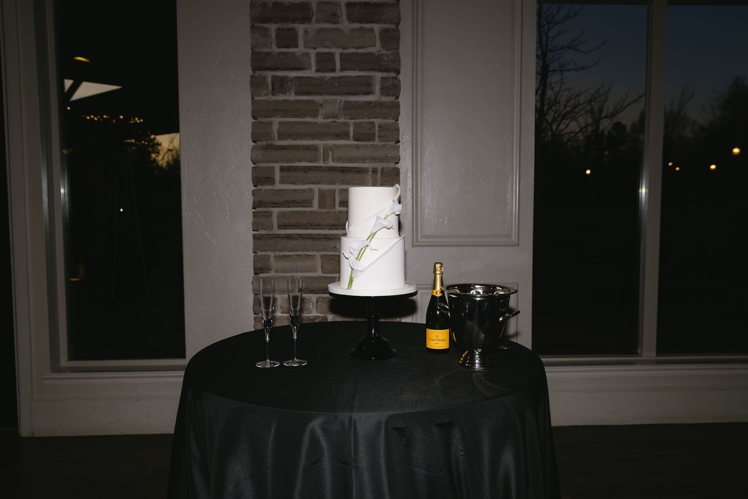 Wedding cake and champagne display on black tablecloth in dimly lit room.