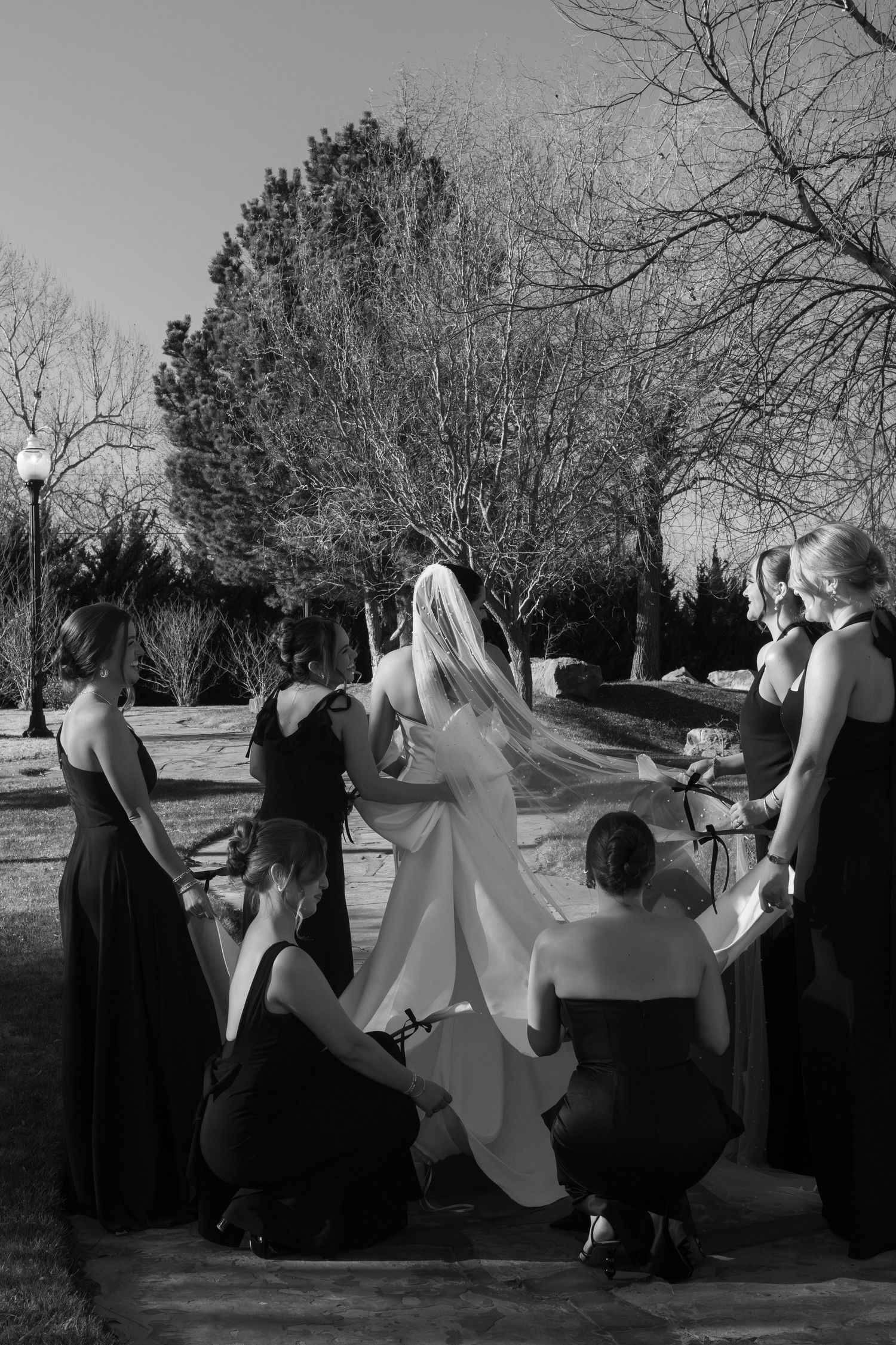 Wedding party in black dresses helping with the bride's dress outdoors.