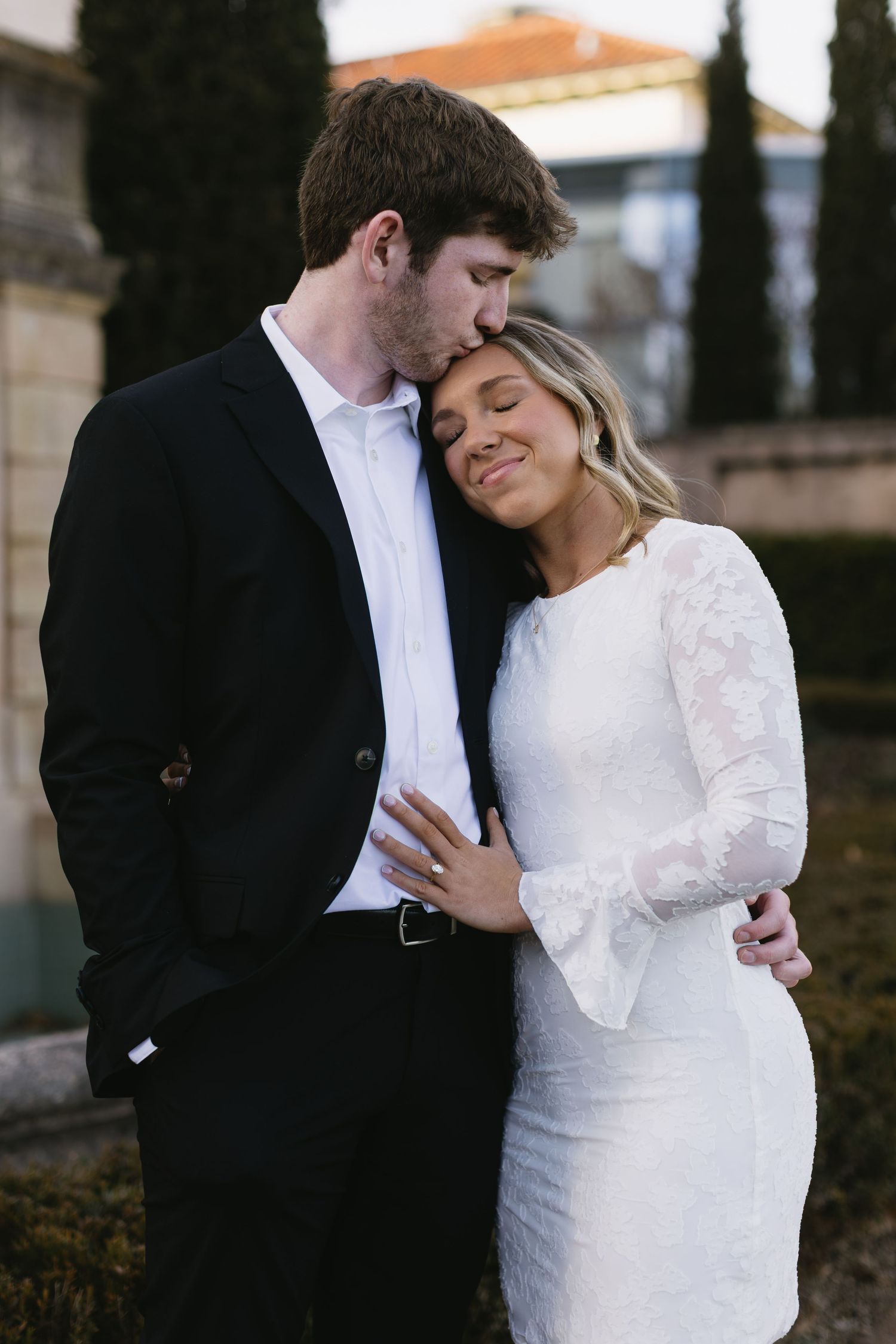 A tender embrace between a couple in formal wear standing near cypress trees and classical architecture.