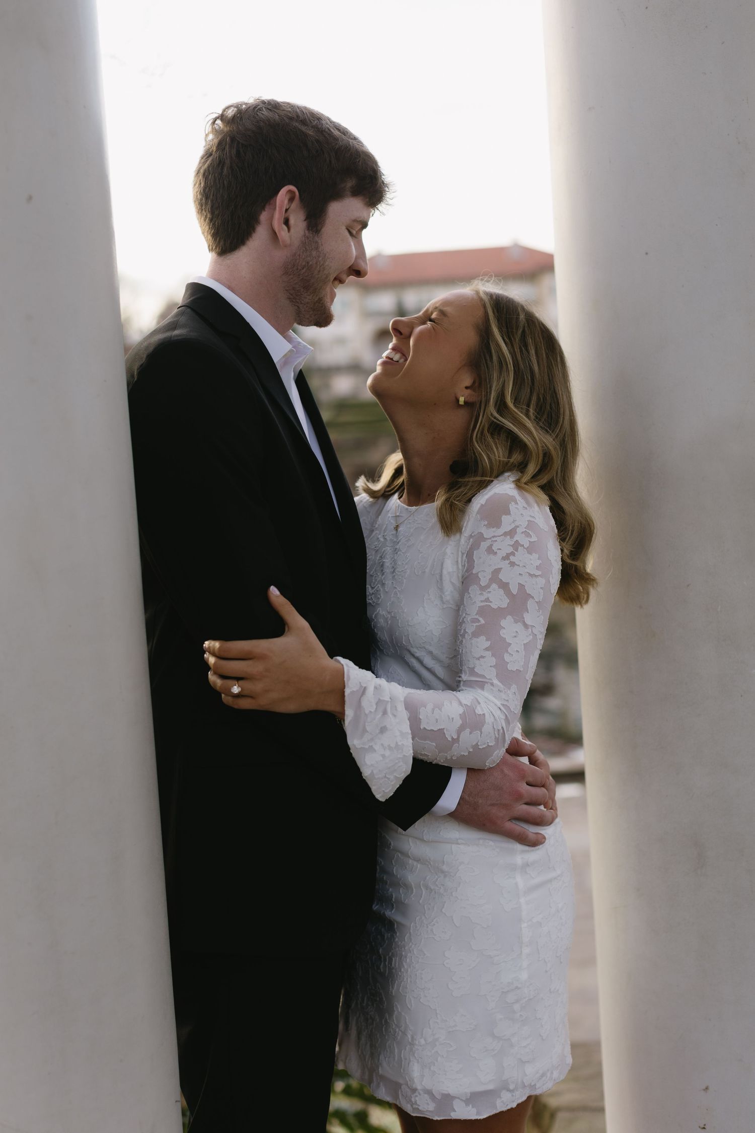 A couple shares a tender moment between white pillars at sunset.
