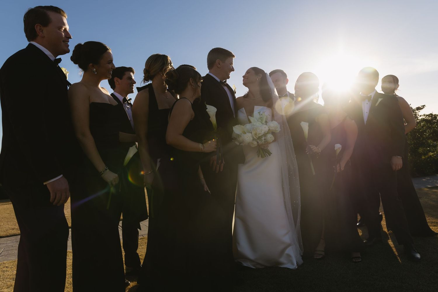 Wedding party silhouetted against bright sunset sky outdoors.