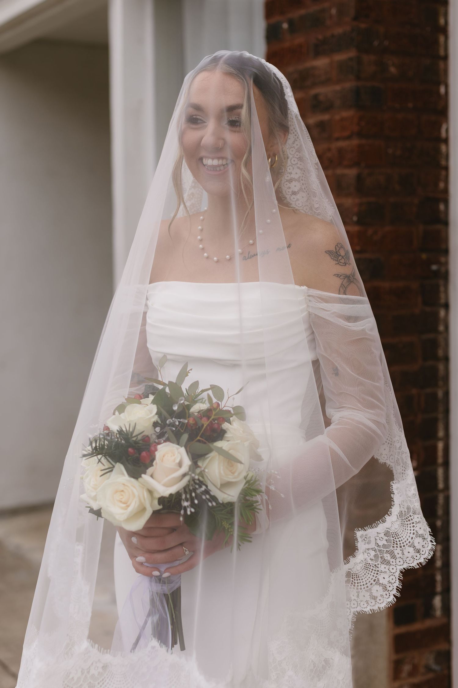 Elegant bride with lace veil holding winter wedding bouquet against brick backdrop.