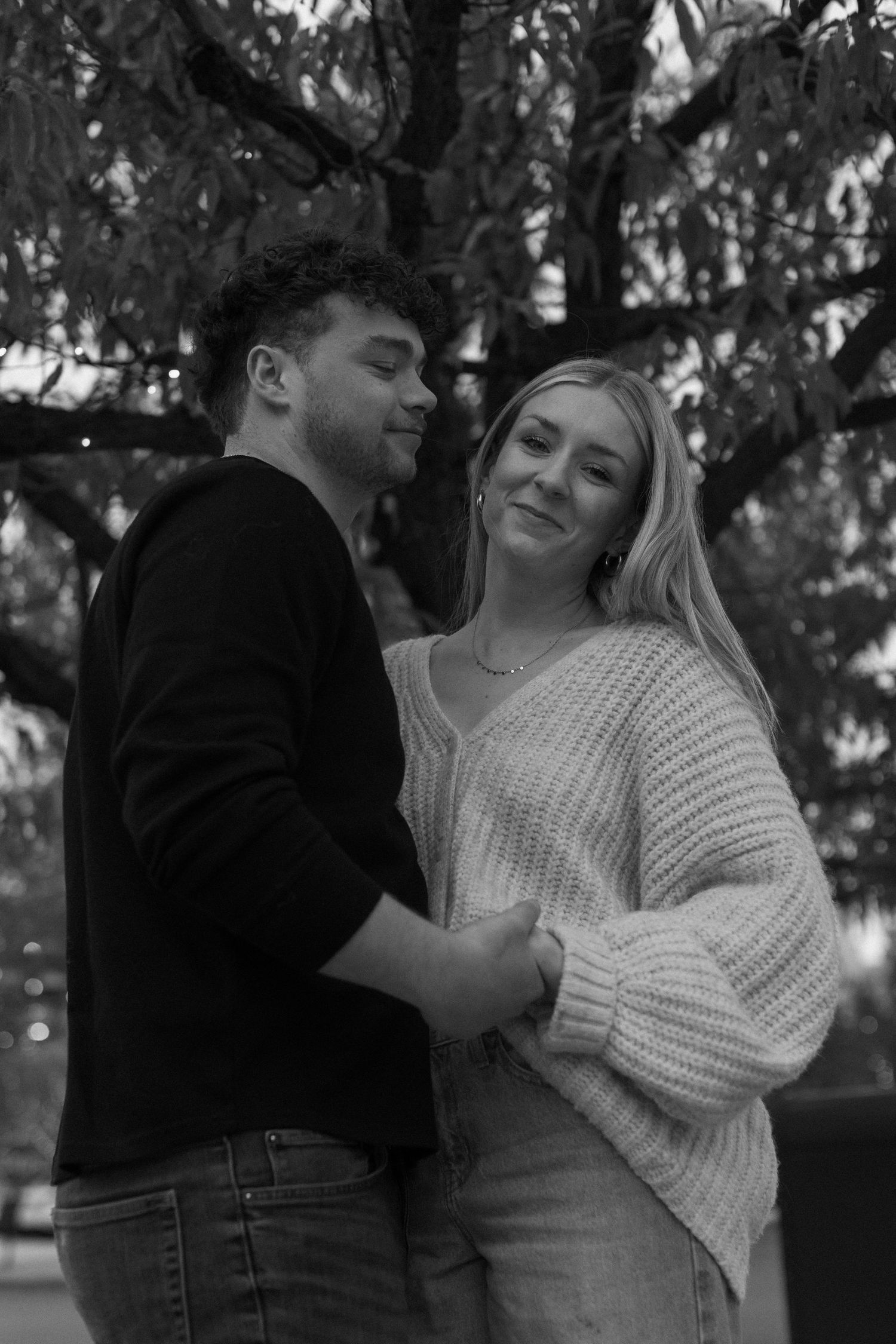 A black and white portrait of a couple embracing outdoors wearing cozy sweaters against a natural backdrop.