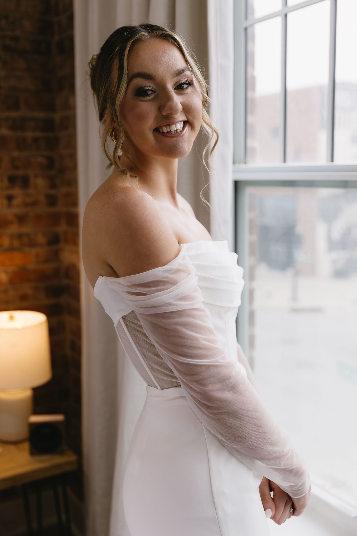 A bride in an elegant off-shoulder white wedding dress with sheer sleeves poses by a window with exposed brick.