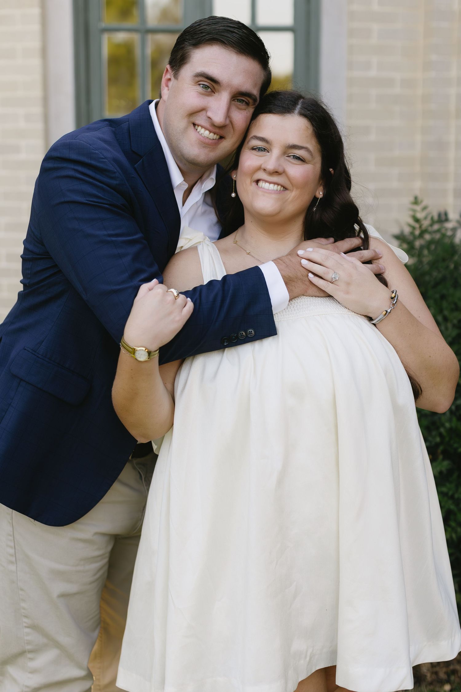 Couple in navy blazer and white dress share intimate moments against beige building exterior.