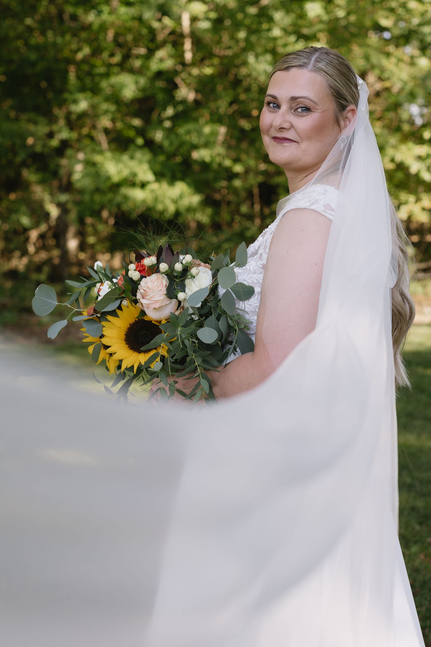 Bride in a flowing white dress holding a sunflower bouquet poses outdoors in natural lighting.