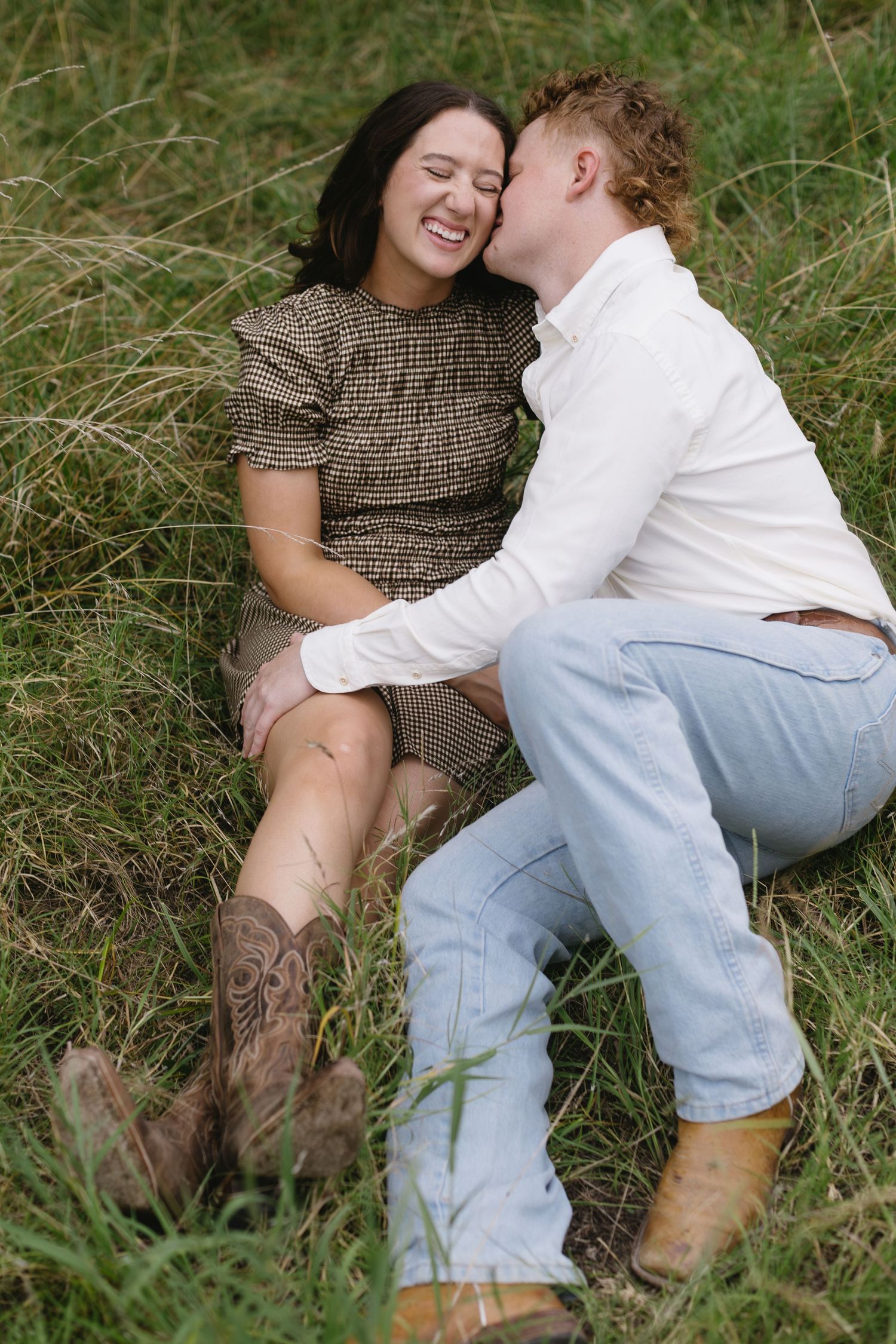 A couple in casual attire lies together in tall grass sharing an intimate moment.