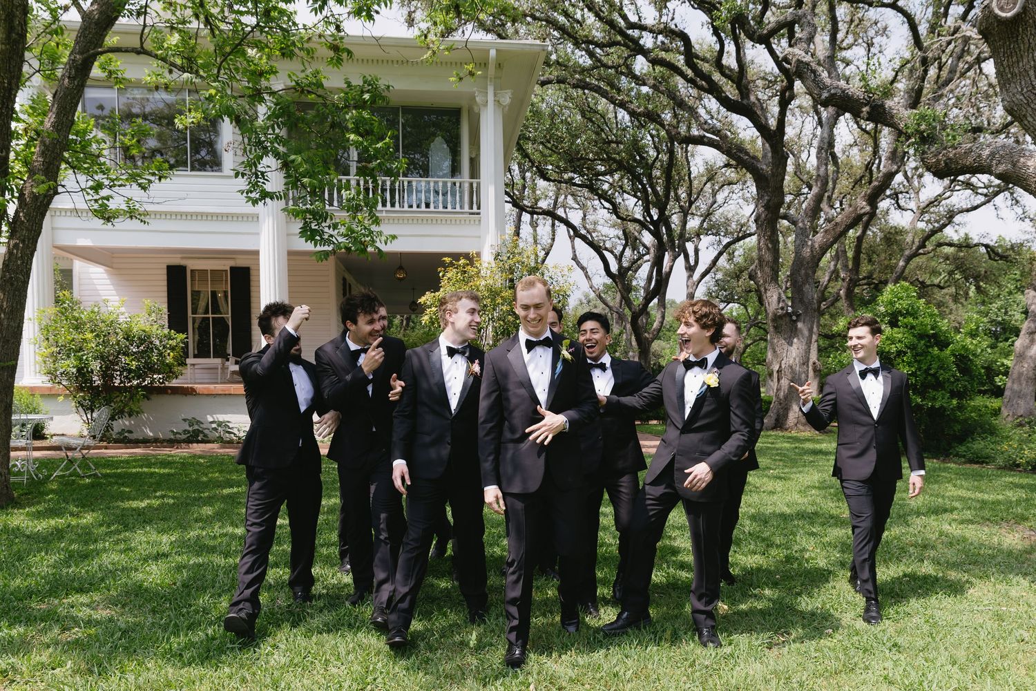 Groomsmen in black tuxedos pose on the lawn in front of a classic southern mansion.
