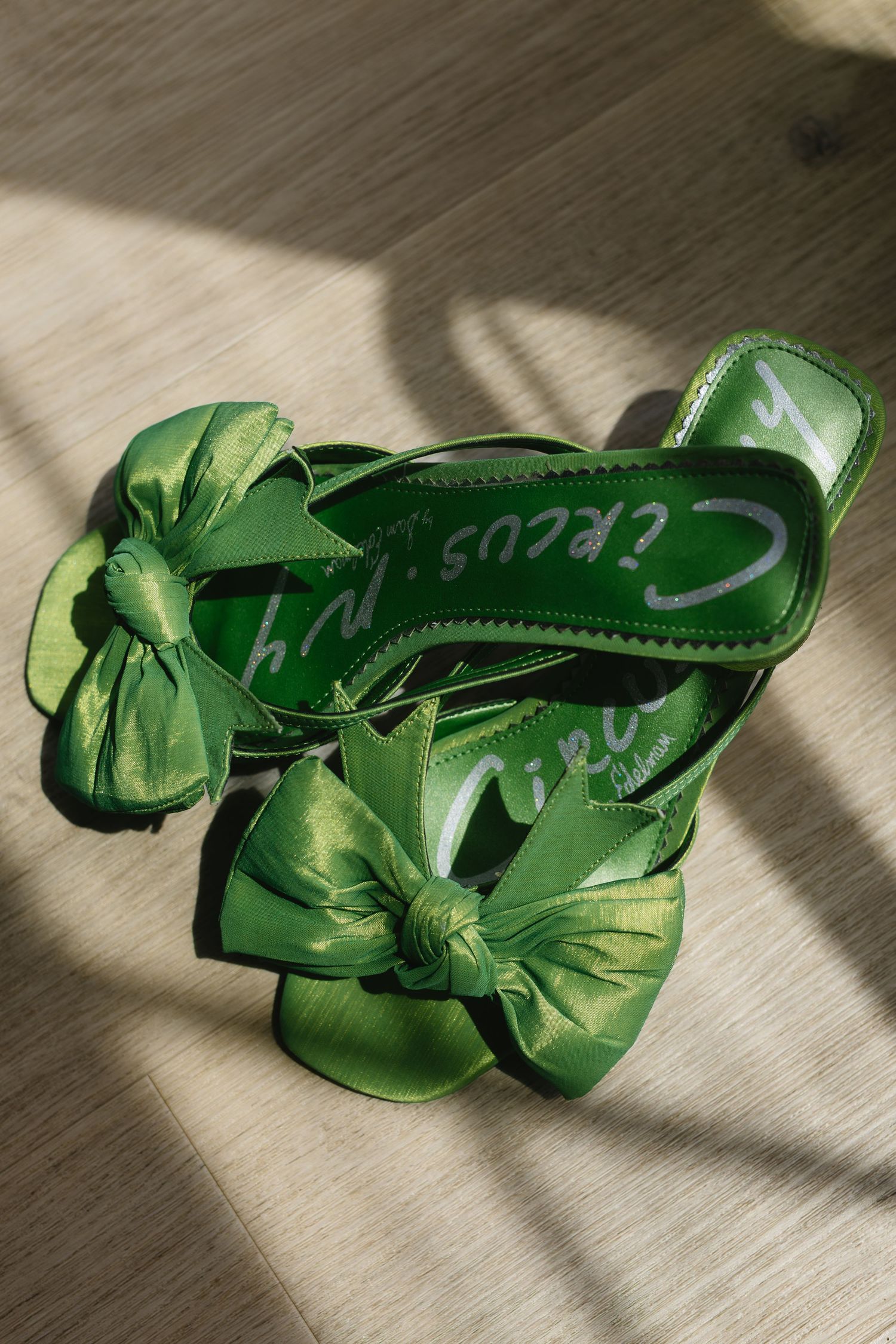 Bright green satin high heel sandals with decorative bows displayed on a light floor with dramatic shadows.