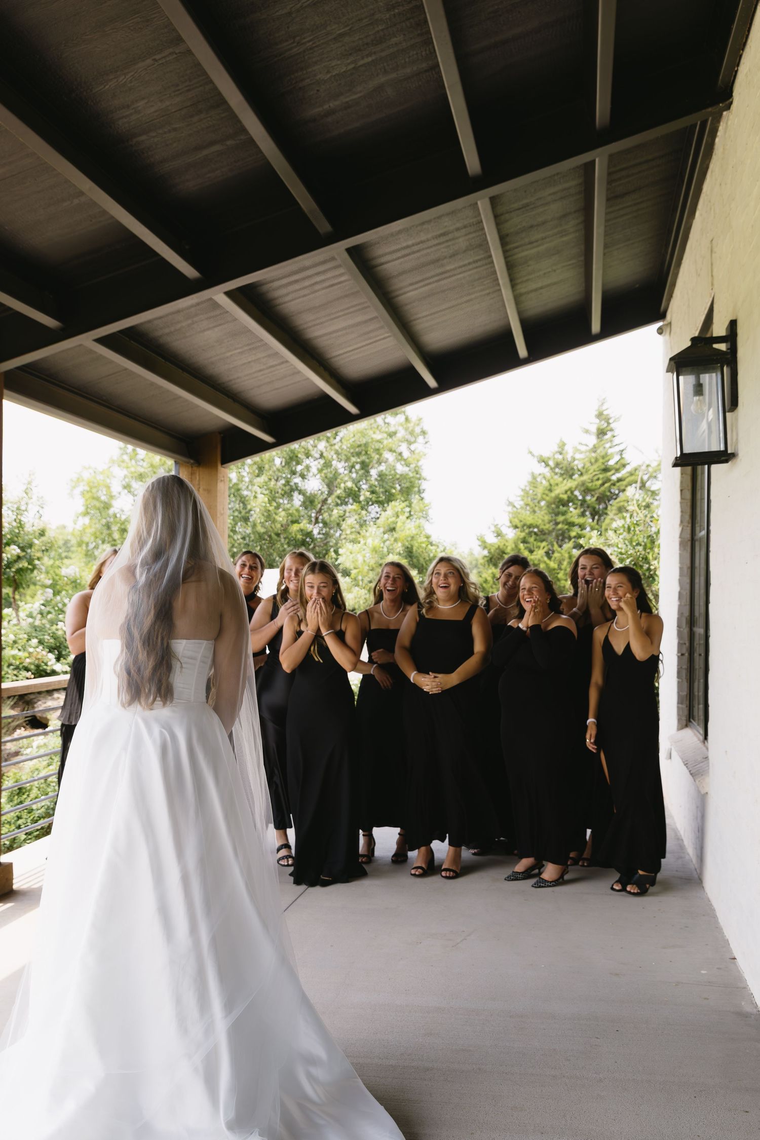 Wedding party in black dresses standing on covered porch with bride in white.