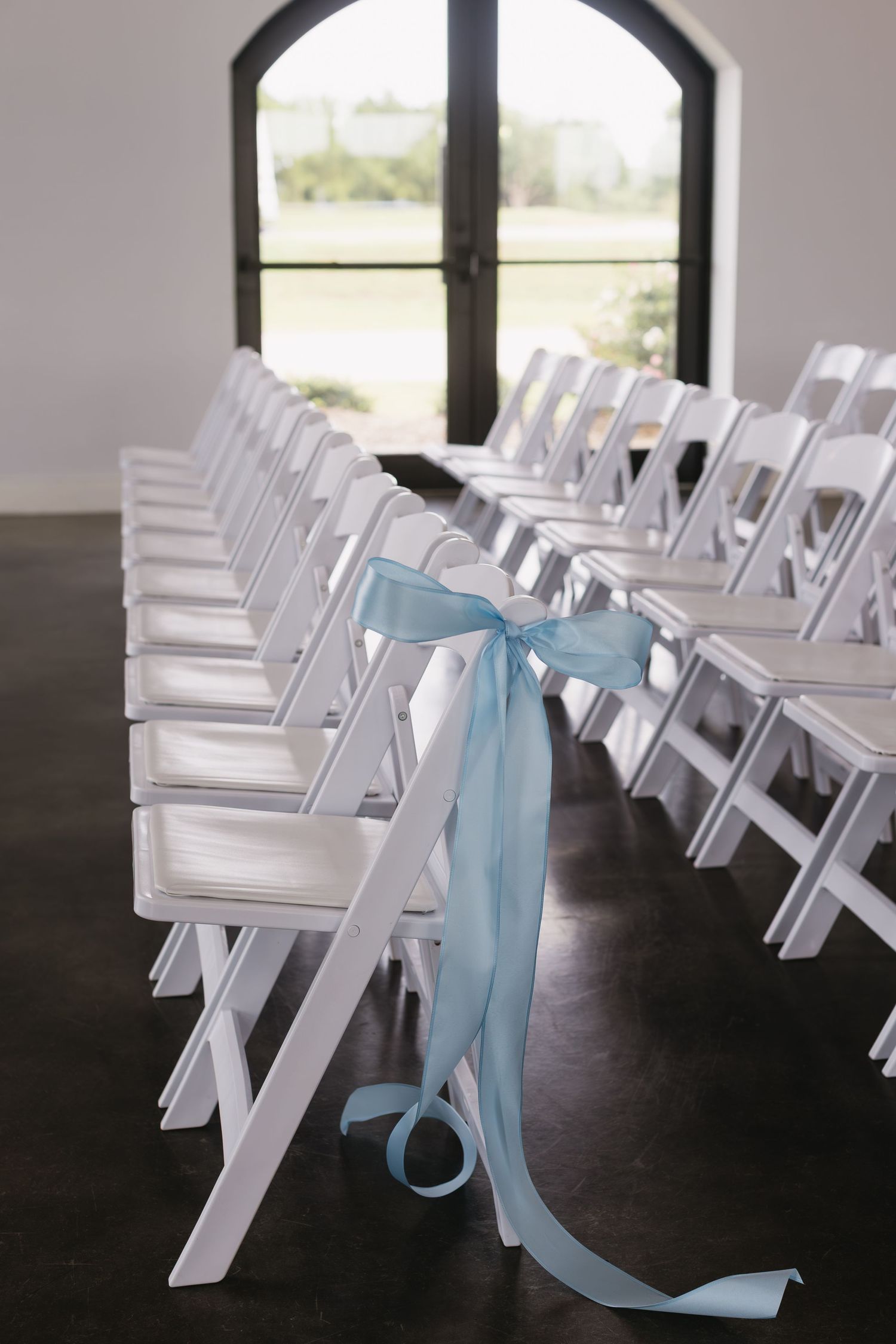 White wedding ceremony chairs arranged in rows with light blue ribbon decorations tied to the ends.