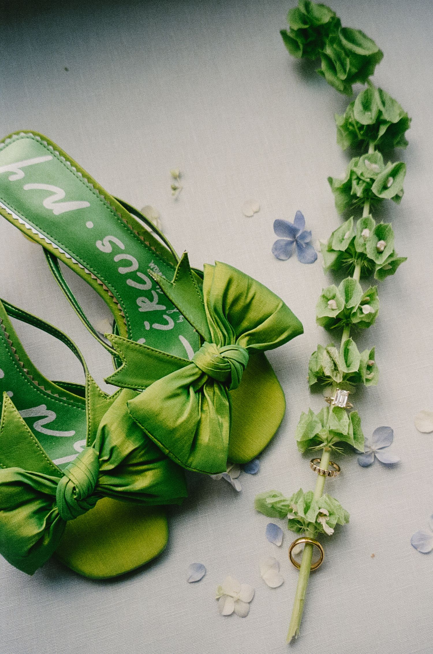 Green bridal shoes with bows displayed alongside scattered blue flower petals and greenery.