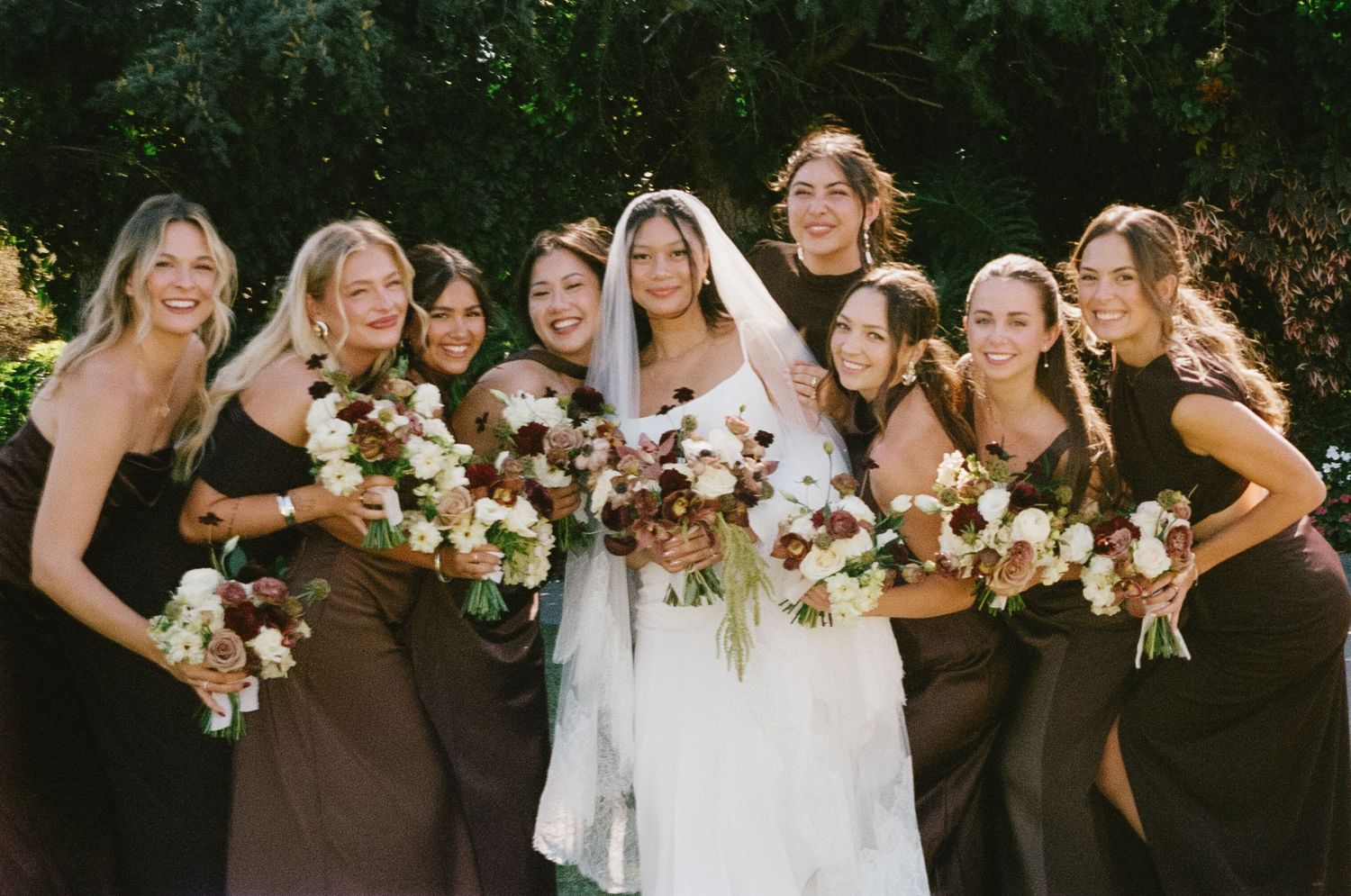 Wedding party in brown dresses and white gown pose together with floral bouquets in an outdoor garden setting.