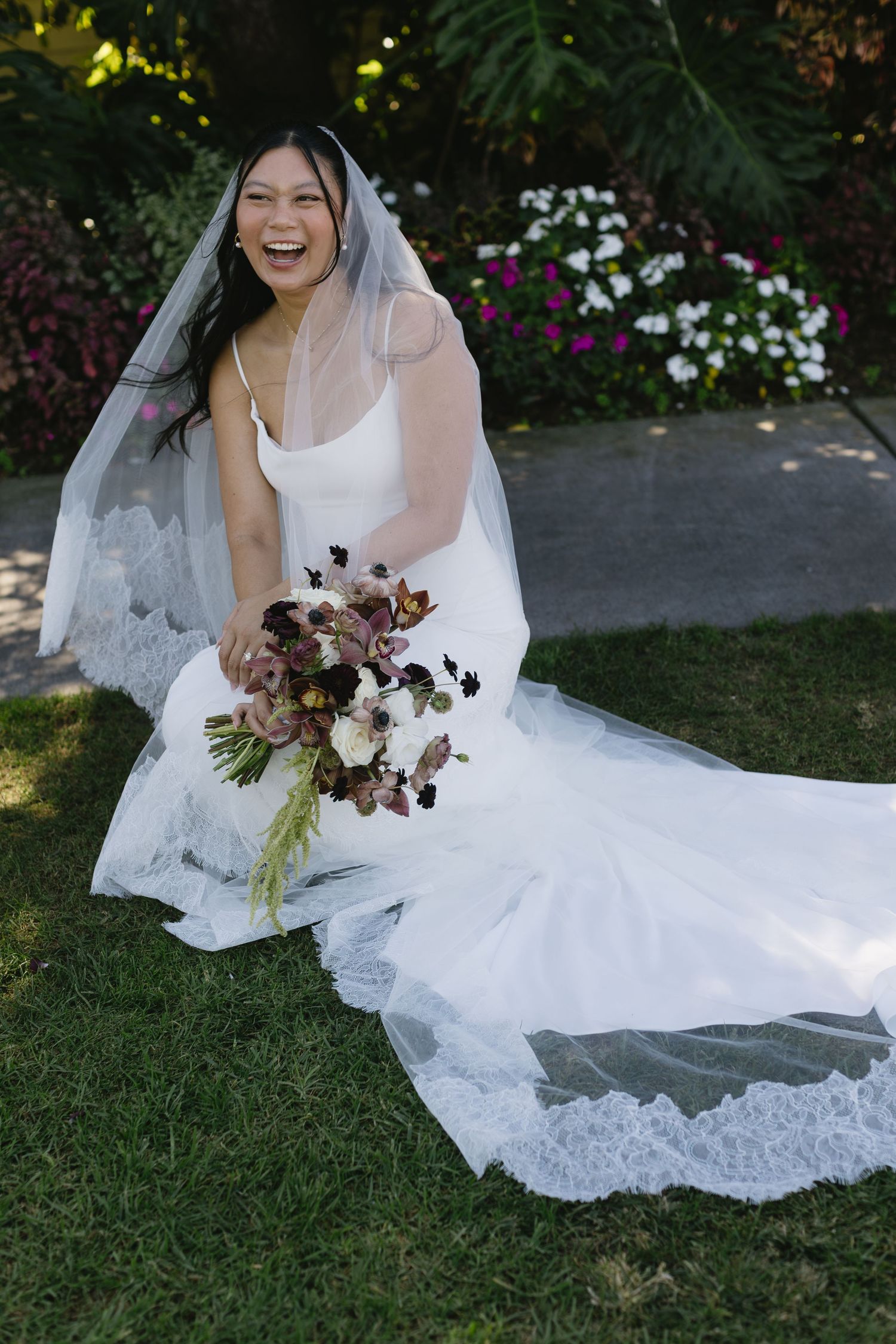 Bride in white lace dress sitting on grass with flowing veil and burgundy bouquet.
