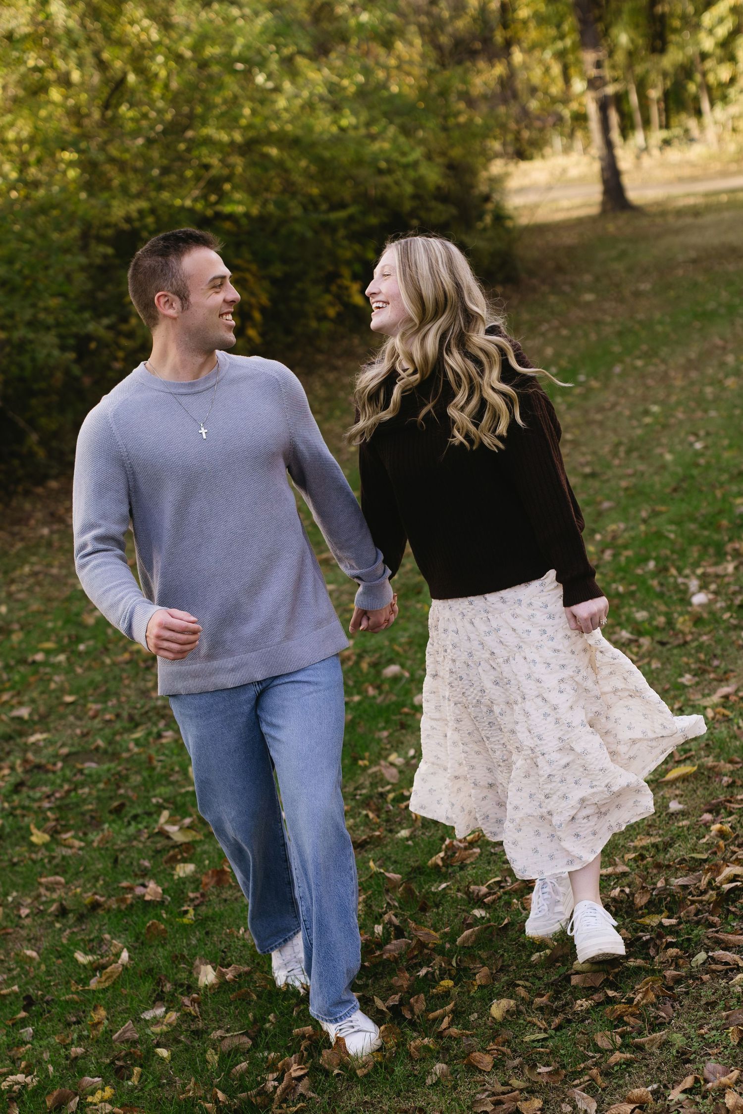 A couple laughing together while walking through fallen autumn leaves in a park.