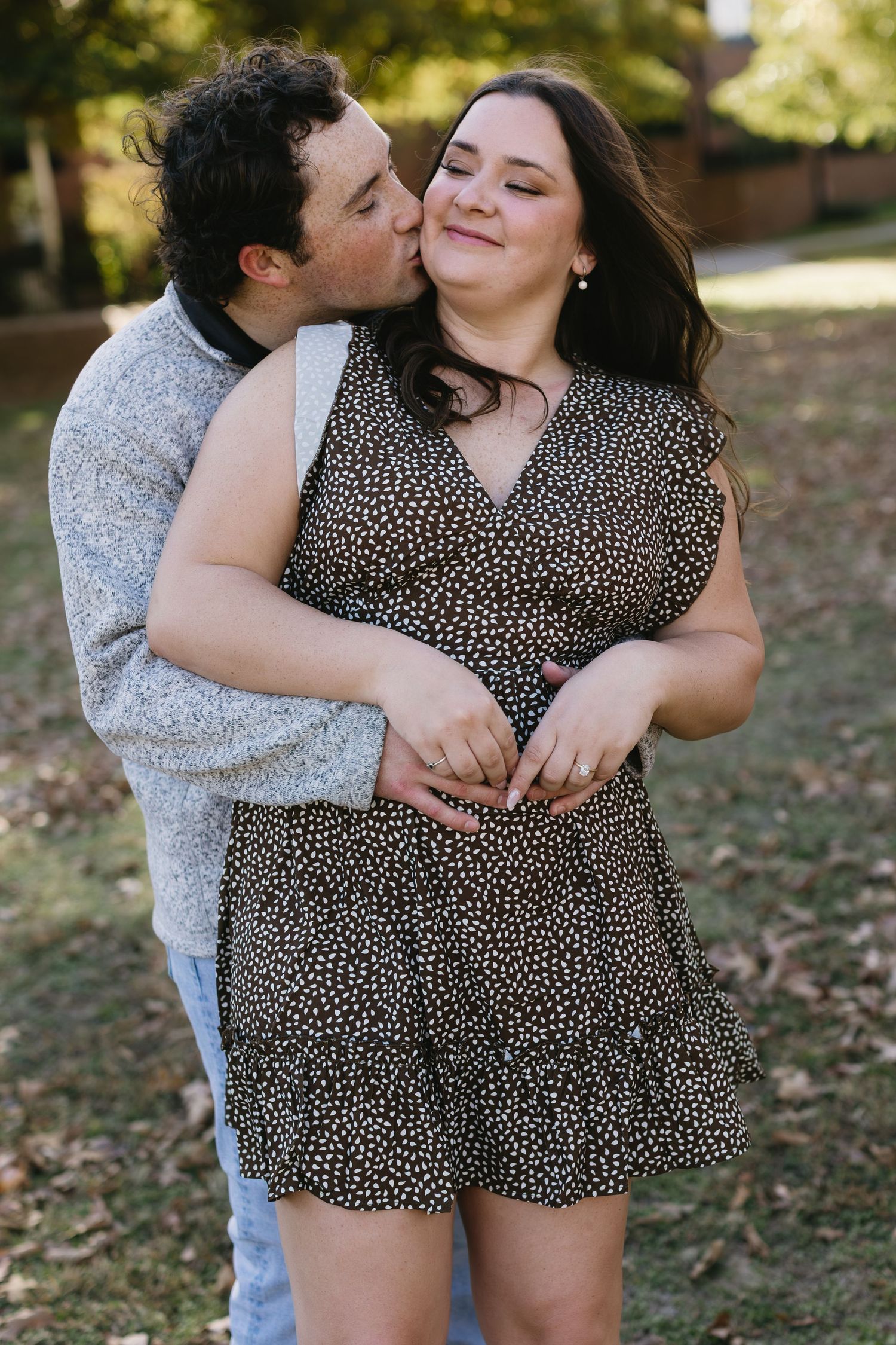 A tender moment between a couple embracing outdoors with sunlit trees in the background.