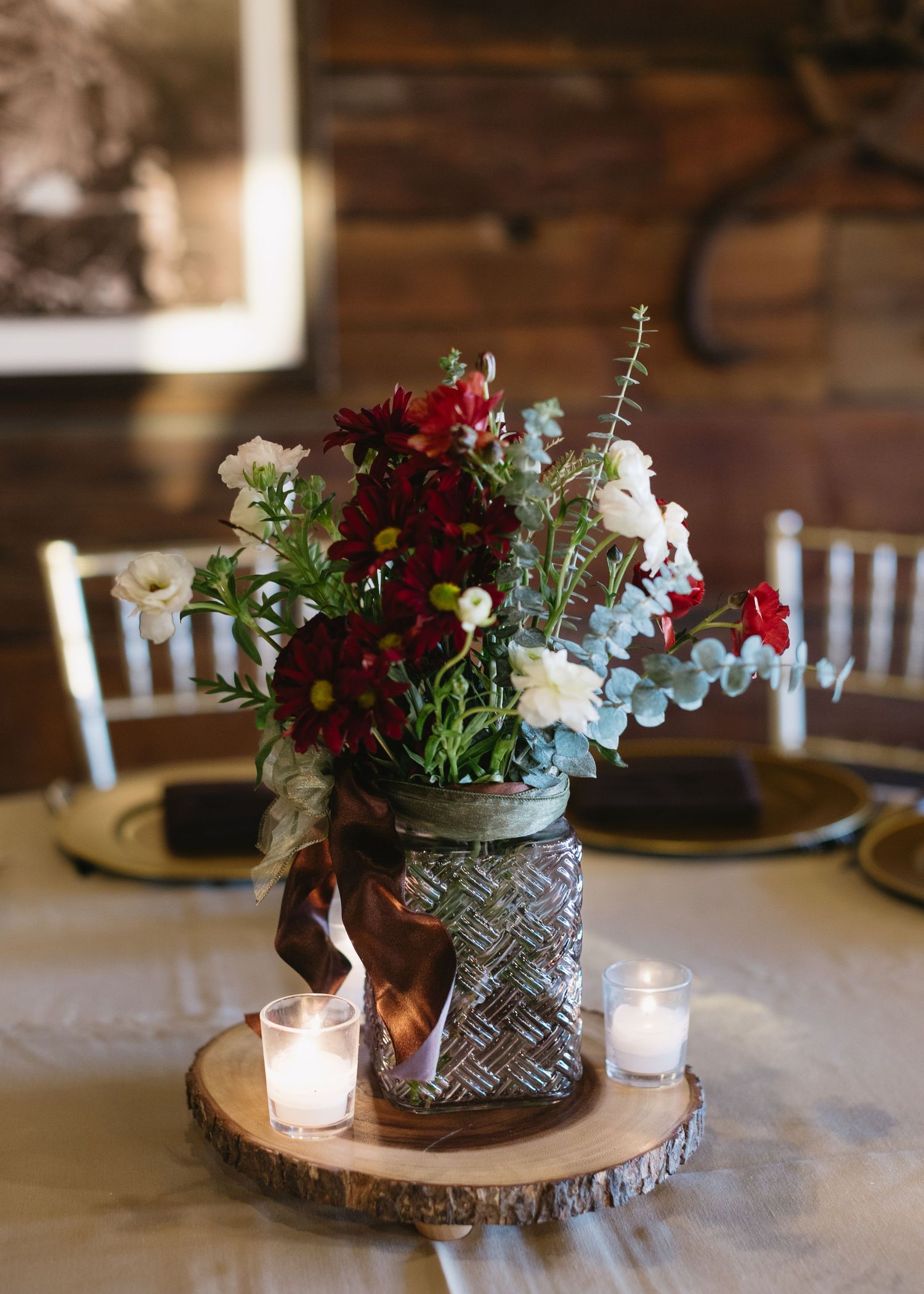 Rustic wedding centerpiece featuring deep red and white flowers arranged in a textured vase on wooden slice charger with candle.