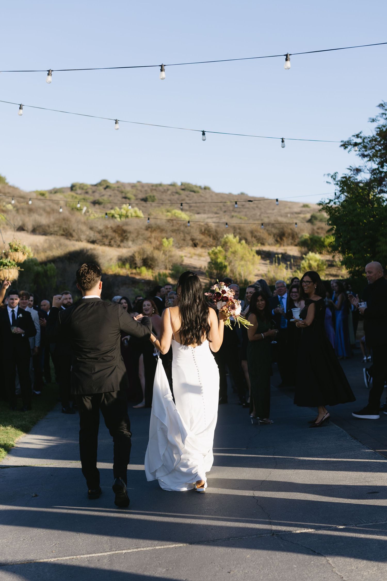 String lights hang above an outdoor wedding reception area as guests gather at sunset in a desert landscape setting.