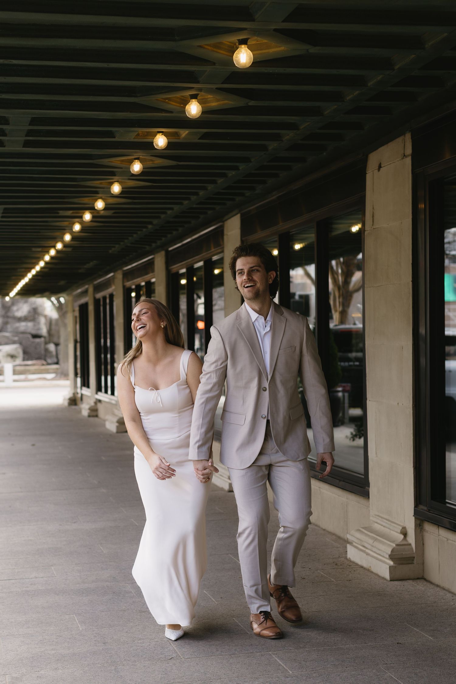 A couple walks hand in hand under a covered walkway with string lights overhead.