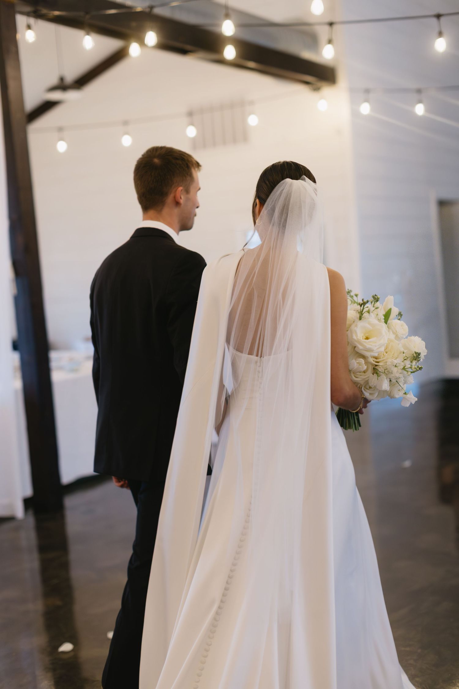 Couple in wedding attire walking together under string lights in an indoor venue.